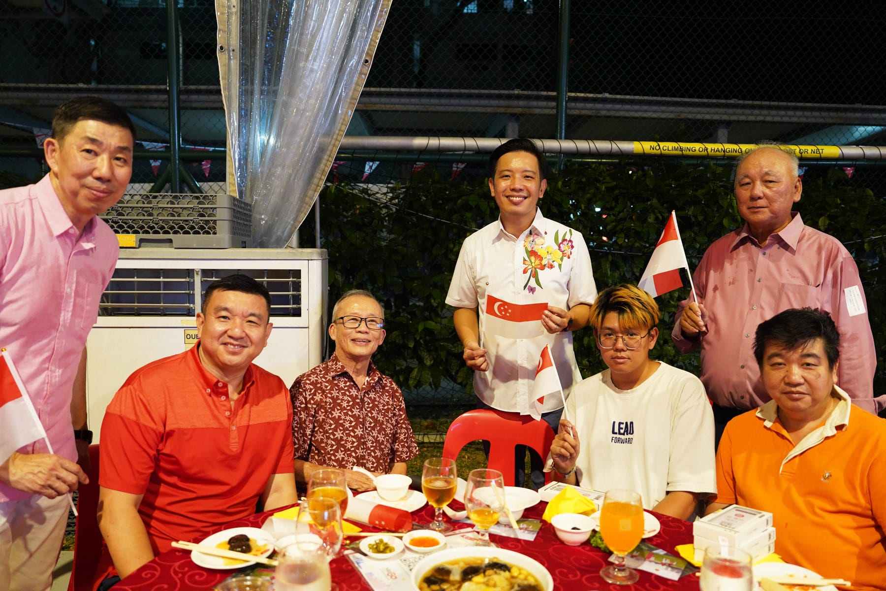 Seven people gathered around a red dining table outdoors at night, holding small Singapore flags and smiling.