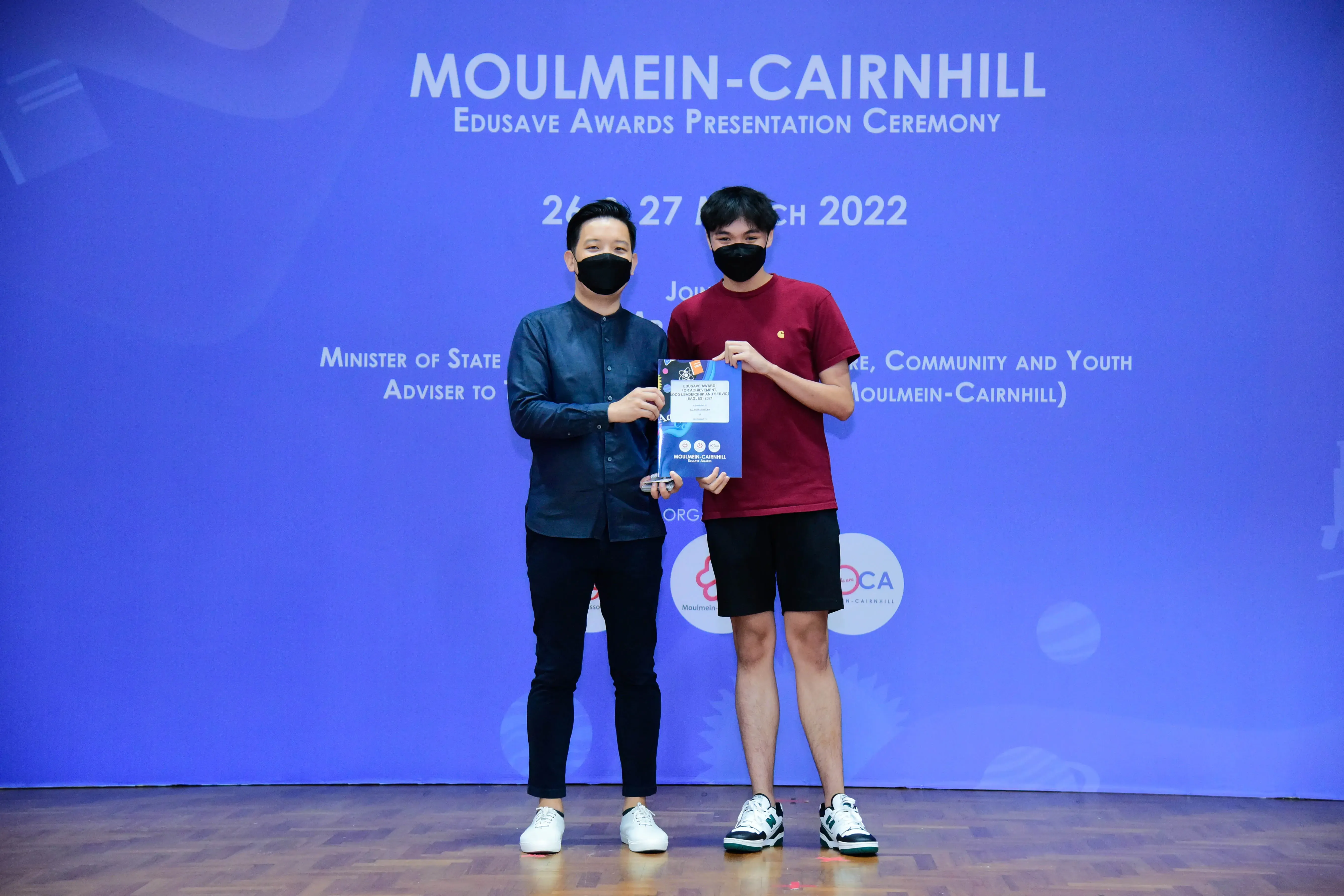 Two masked people hold an award certificate on stage at the Moulmein-Cairnhill Edusave Awards Ceremony 2022.