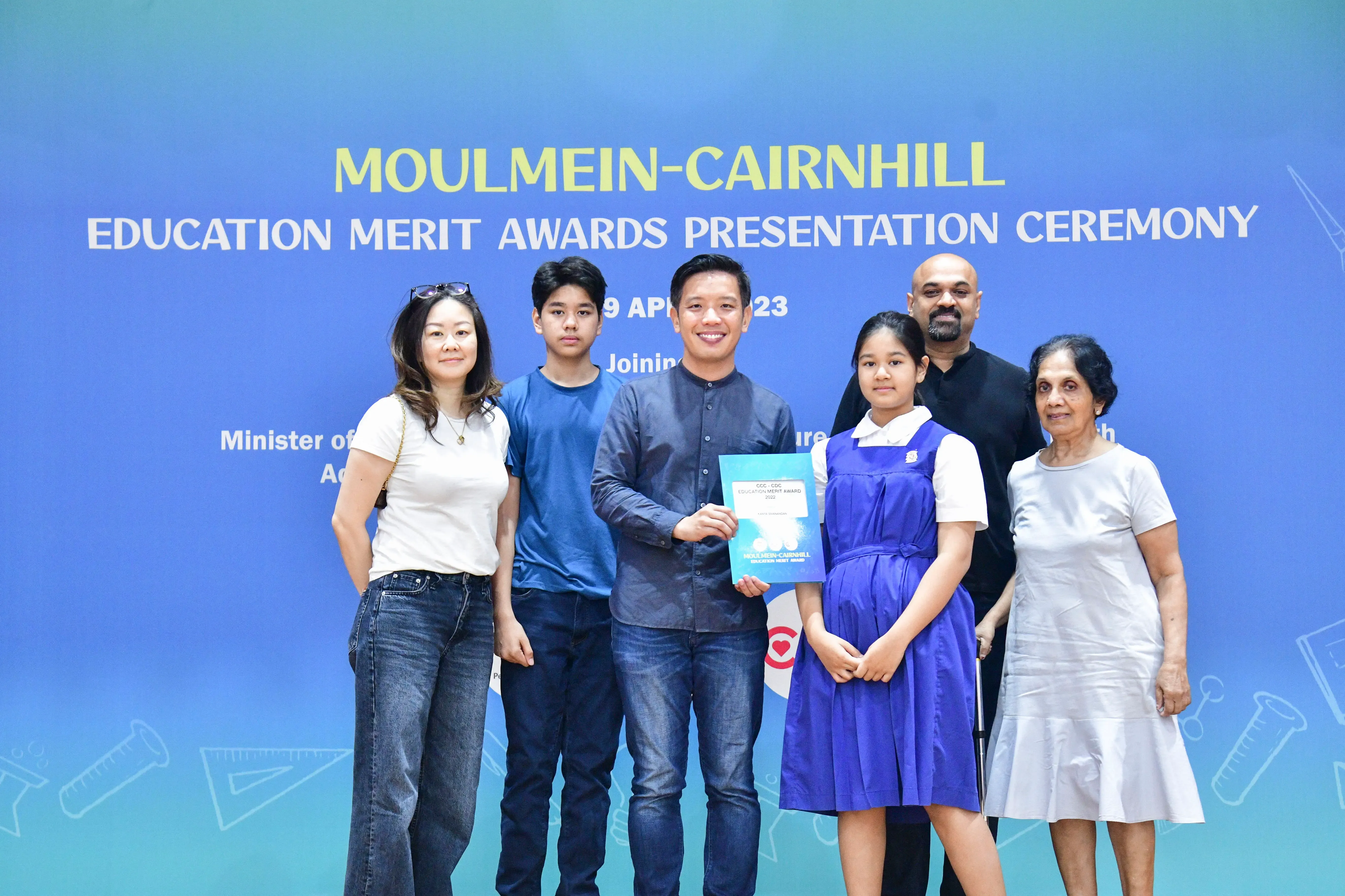 Six people pose in front of a blue banner for the Moulmein-Cairnhill Education Merit Awards Presentation Ceremony.