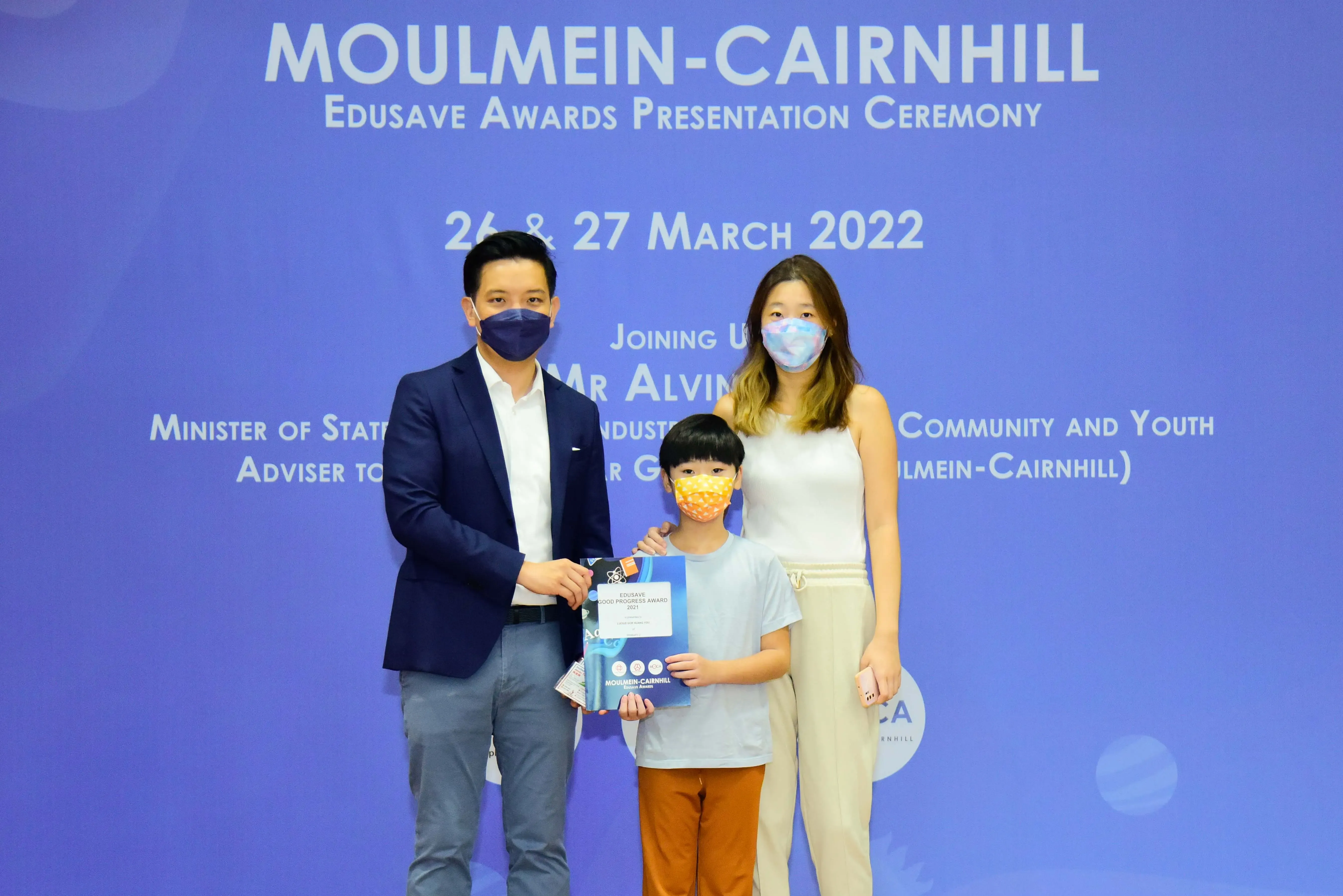 Two masked adults and a masked child hold an Edusave award certificate at the Moulmein-Cairnhill ceremony in March 2022.