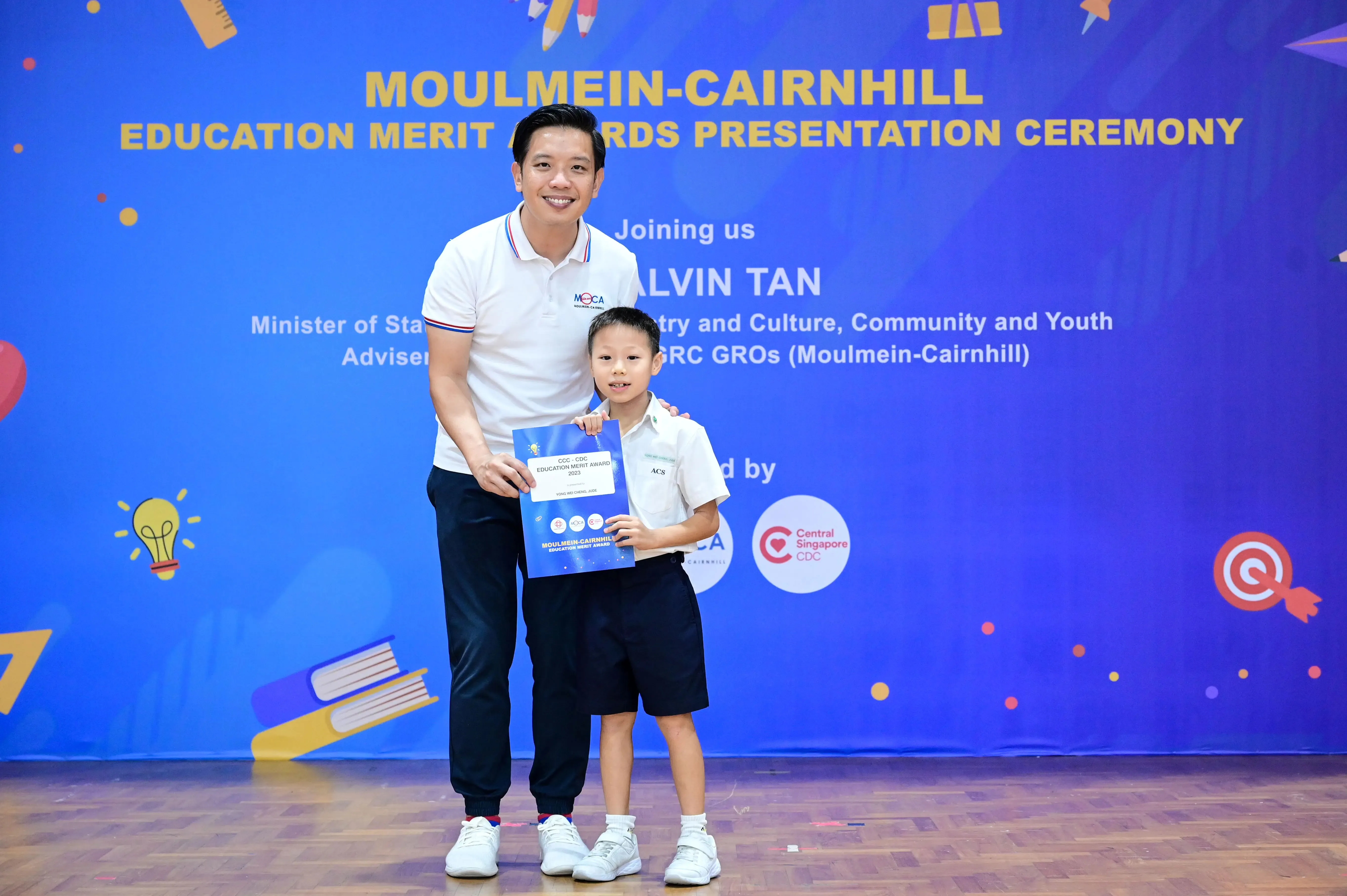 Man and young boy holding a certificate in front of a Moulmein-Cairnhill Education Merit Awards banner.