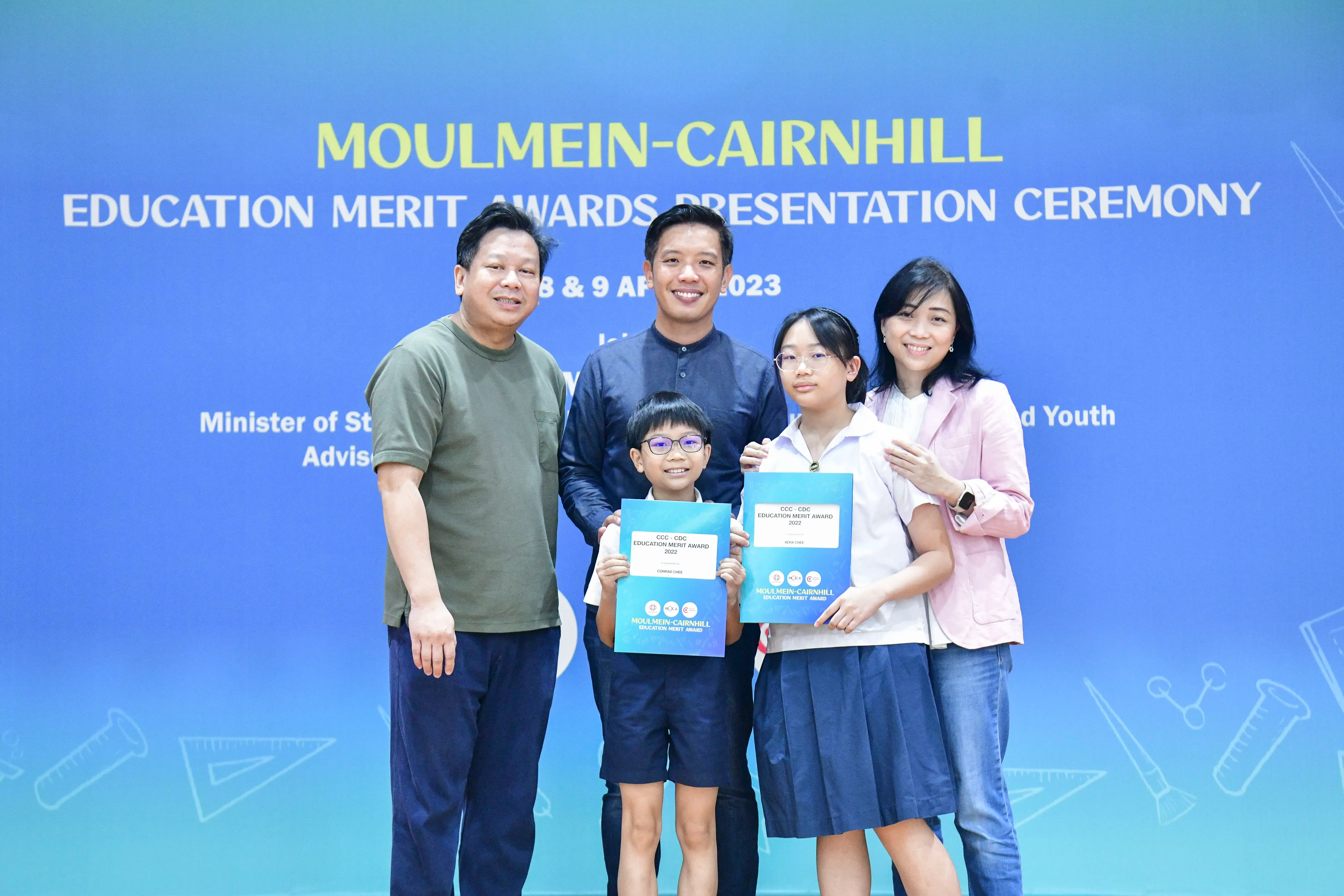 Five people pose before a blue Moulmein-Cairnhill Education Merit Awards banner, two children holding award certificates.