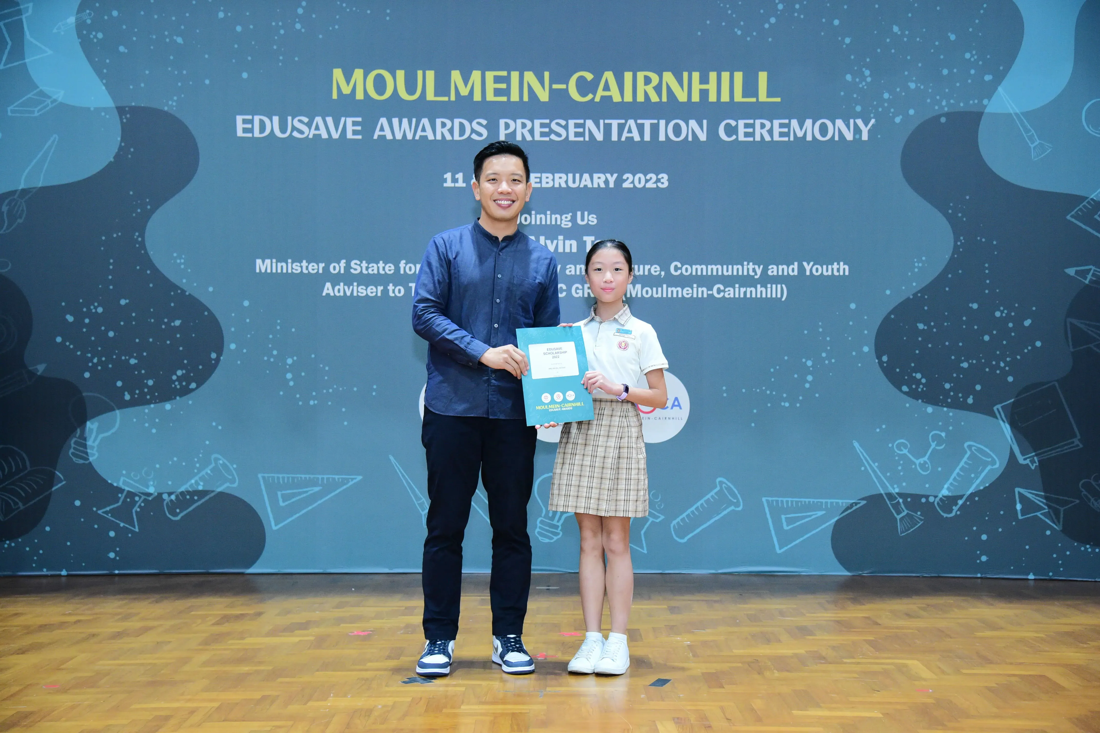 A man and a young girl hold a certificate together on stage at the Moulmein-Cairnhill Edusave Awards Ceremony 2023.