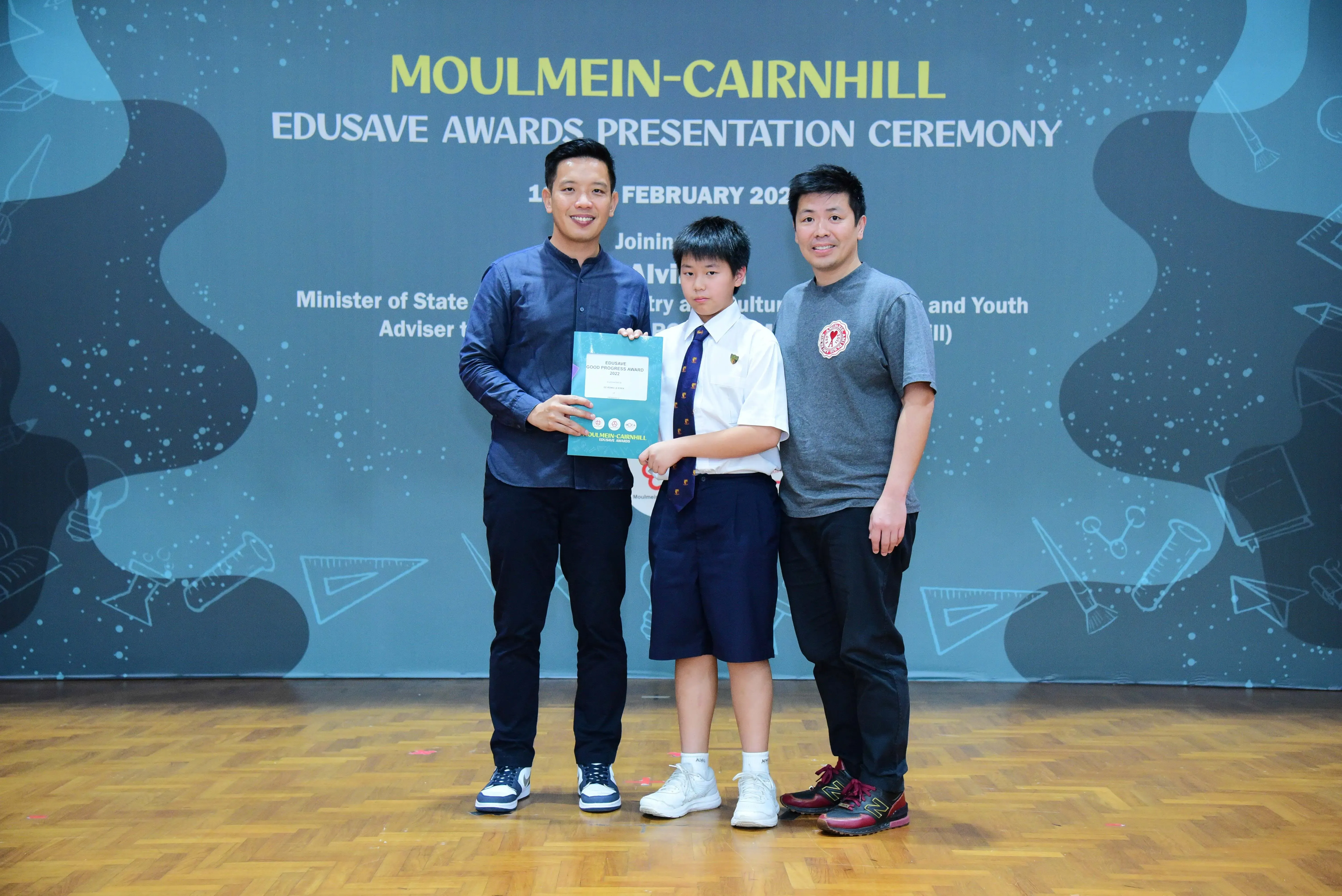 Two men and a uniformed student hold a certificate at the Moulmein-Cairnhill Edusave Awards Presentation Ceremony.