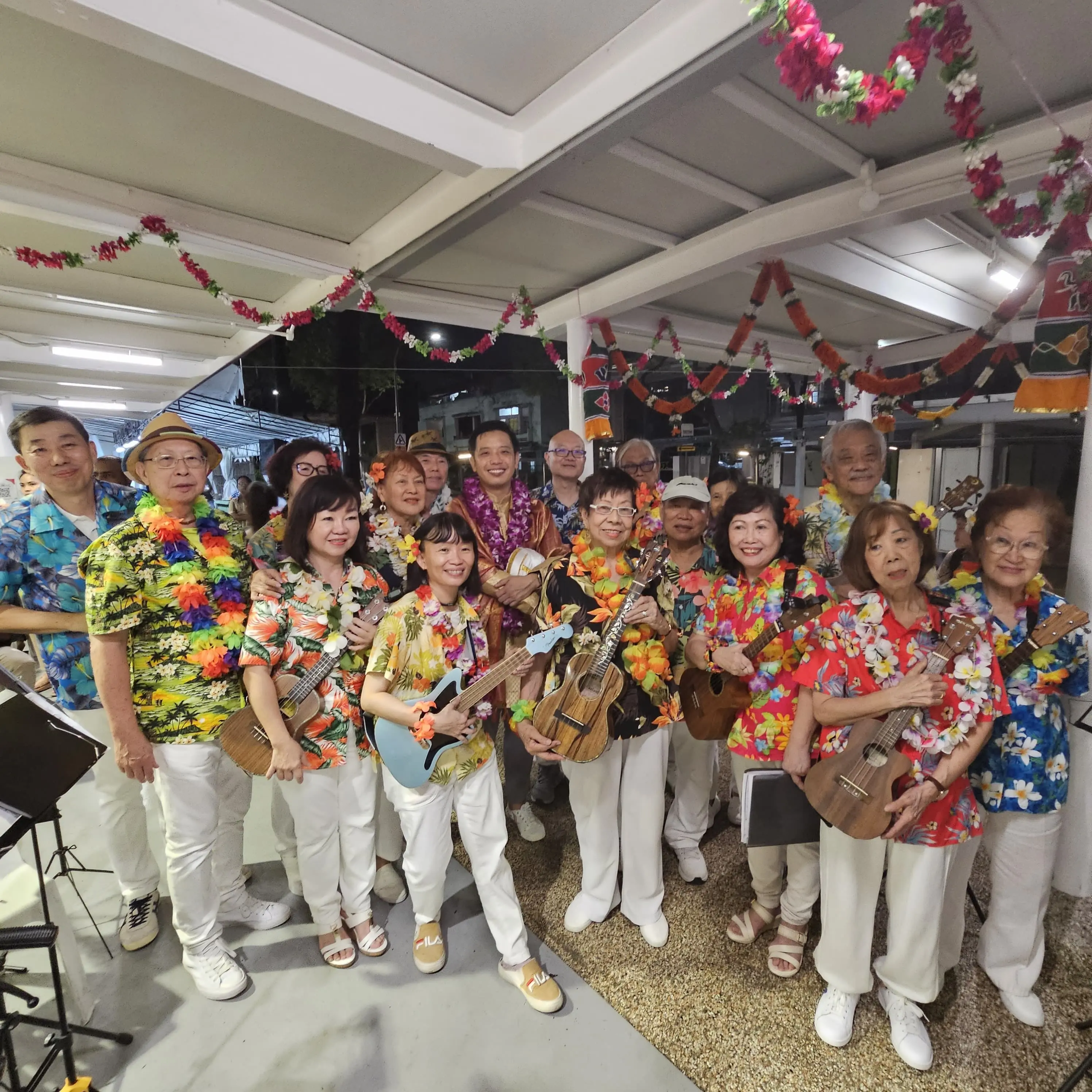 Group of adults in Hawaiian shirts and flower leis holding ukuleles, posing indoors under floral garland decorations.
