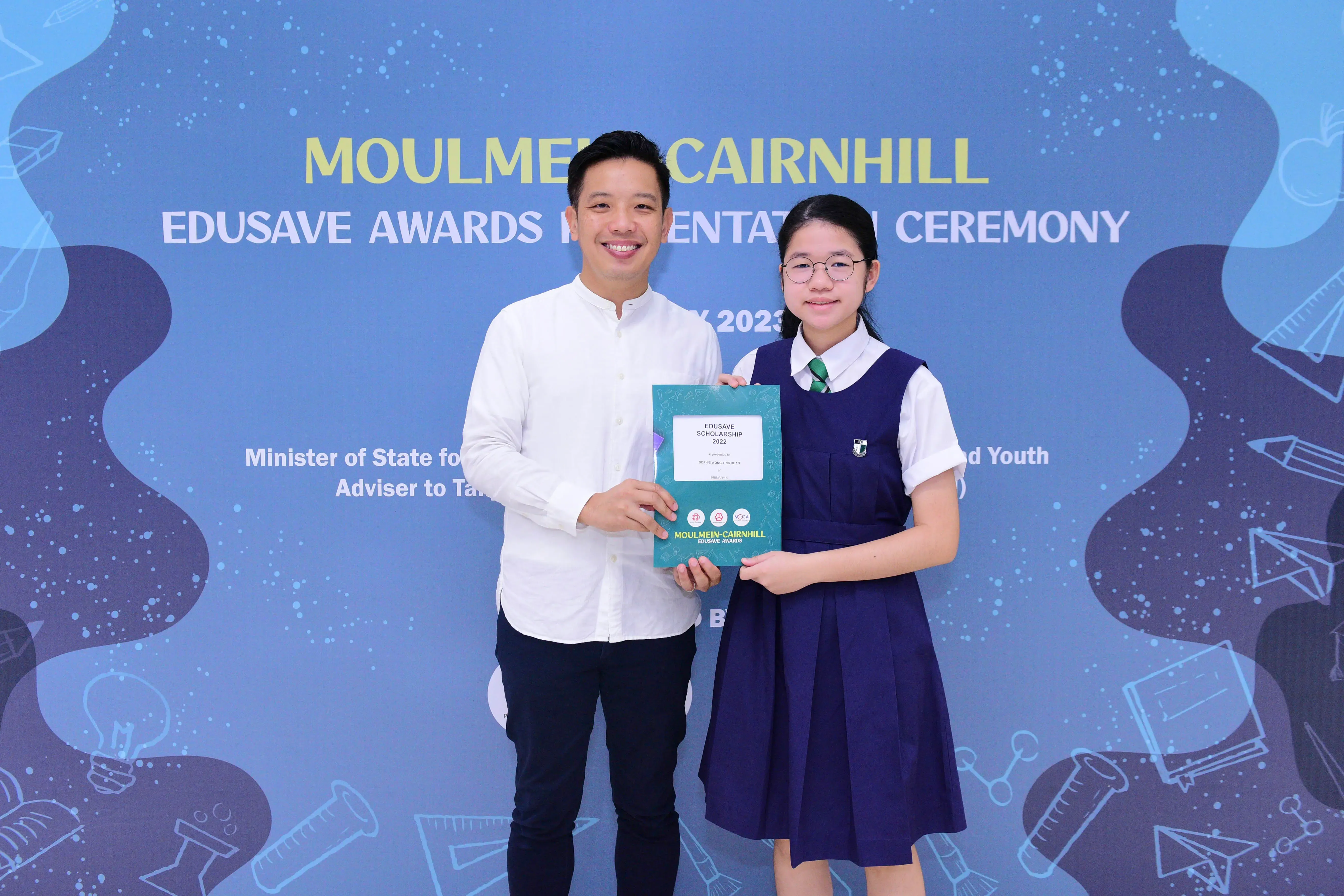 A man and a young girl in school uniform hold an Edusave Scholarship certificate at the Moulmein-Cairnhill Awards Ceremony.