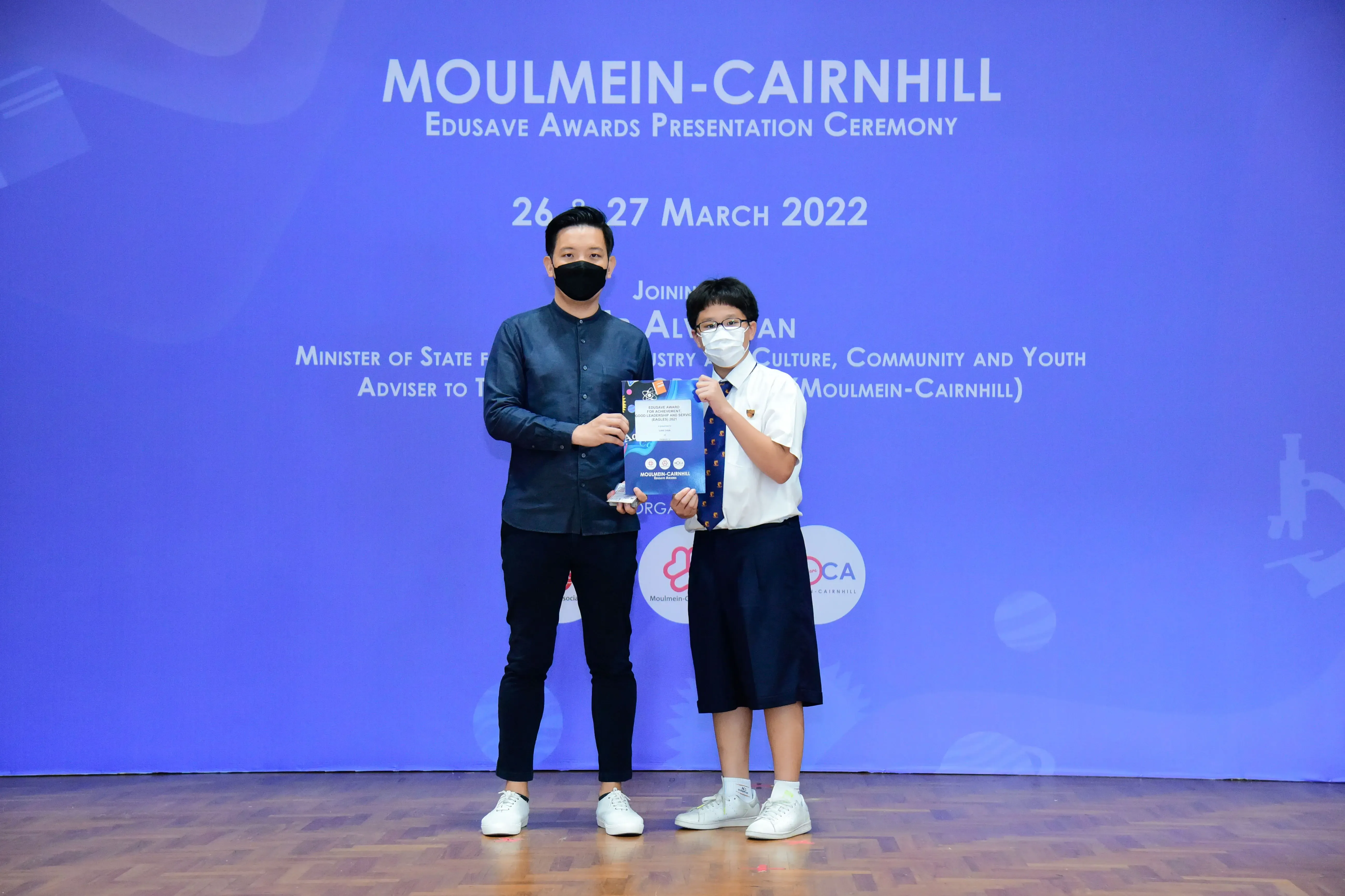 An adult and a masked student hold an award at the Moulmein-Cairnhill Edusave Awards Ceremony, March 2022.