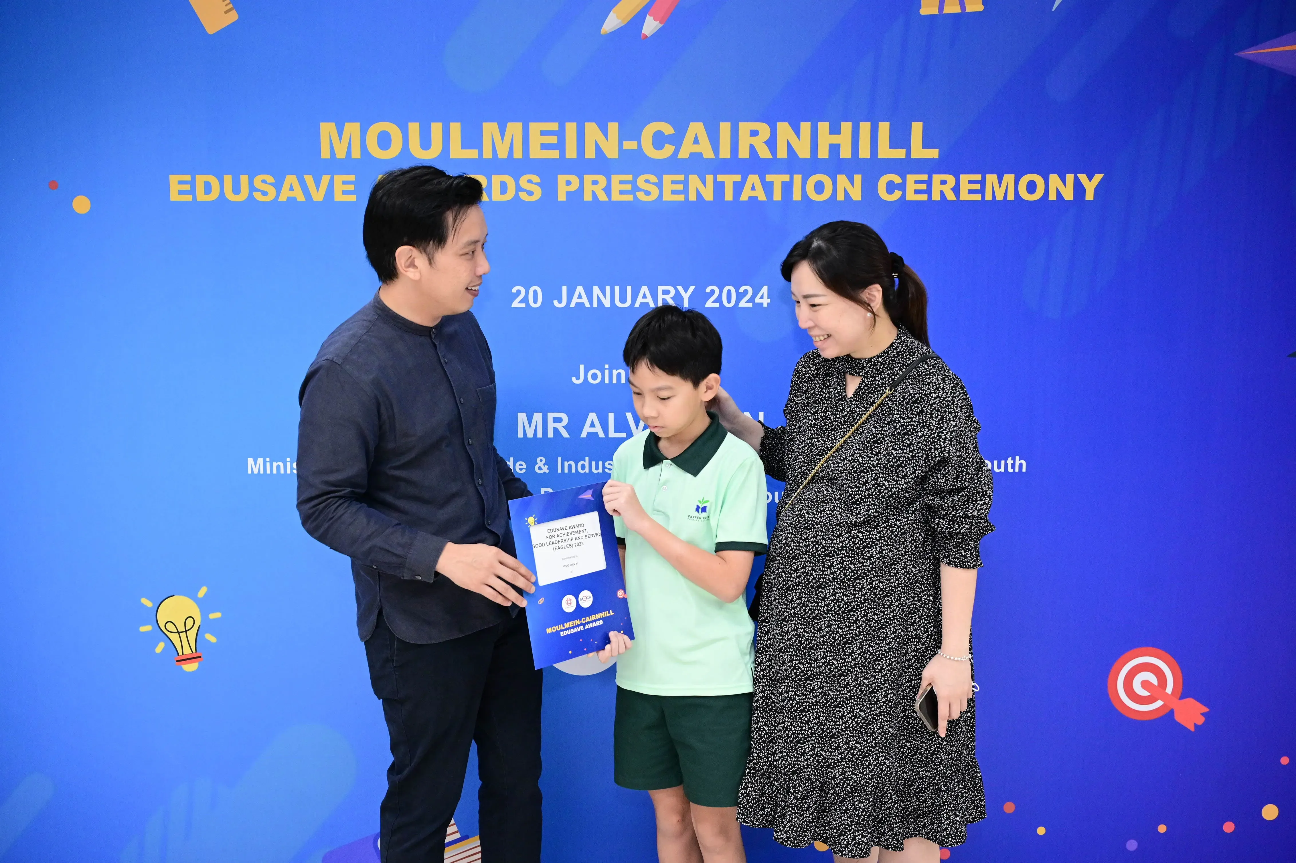 A boy holds a certificate between two adults at the Moulmein-Cairnhill Edusave Awards Presentation Ceremony, January 2024.