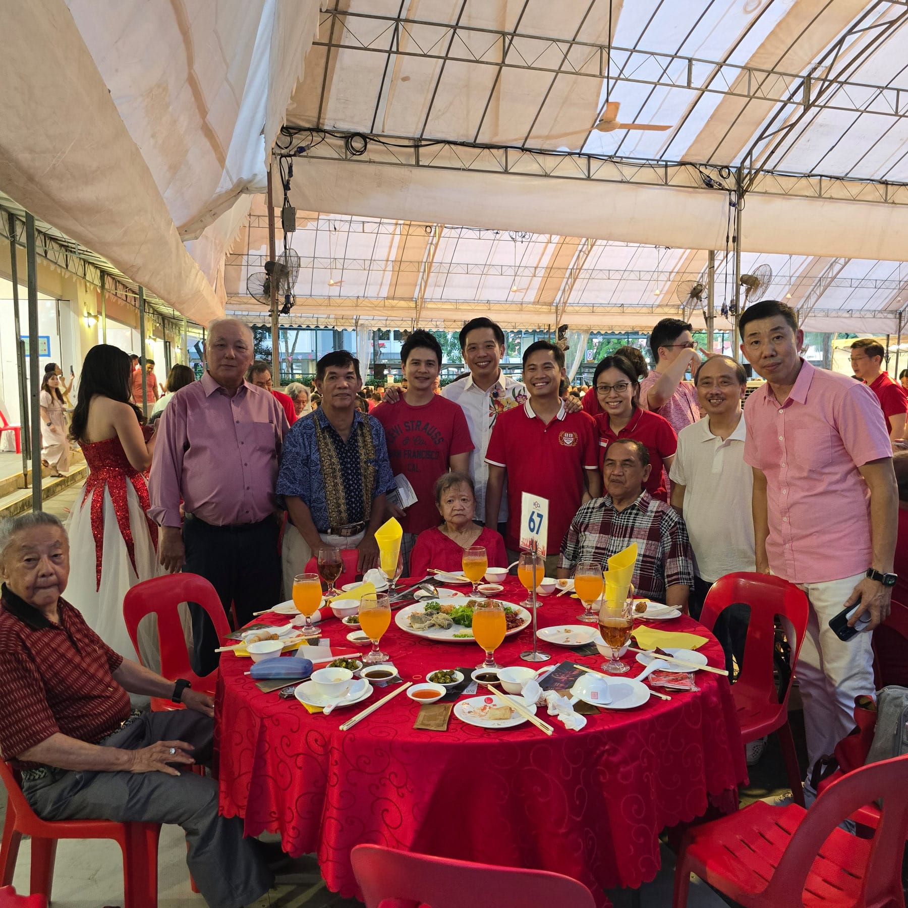 A group of people posing around a red-clothed round dining table with food and drinks at an outdoor banquet event.