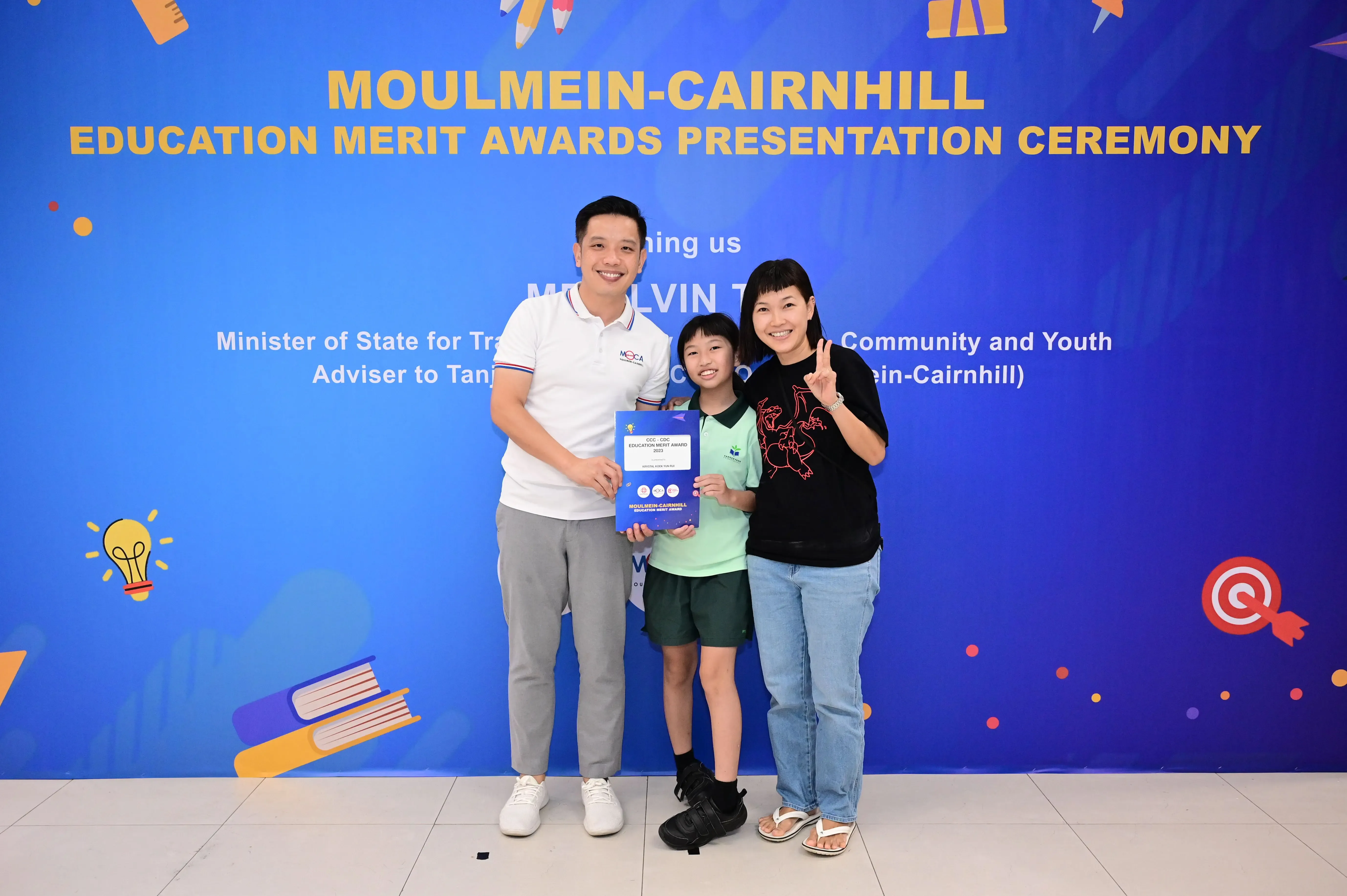 Three people pose smiling before a blue Moulmein-Cairnhill Education Merit Awards banner, with a child holding an award certificate.
