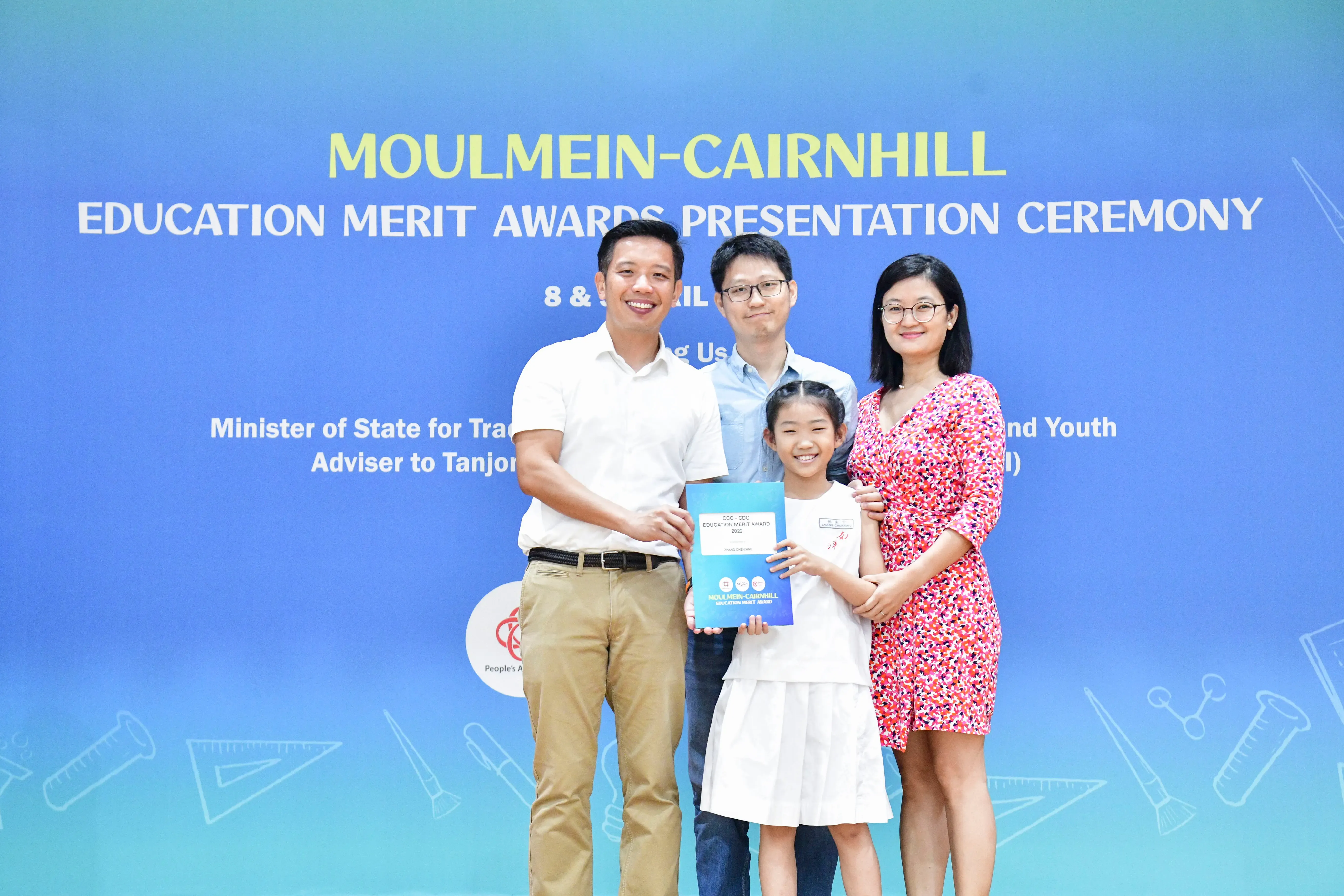 Four people pose before a Moulmein-Cairnhill Education Merit Awards Presentation Ceremony banner, a girl holds an award folder.