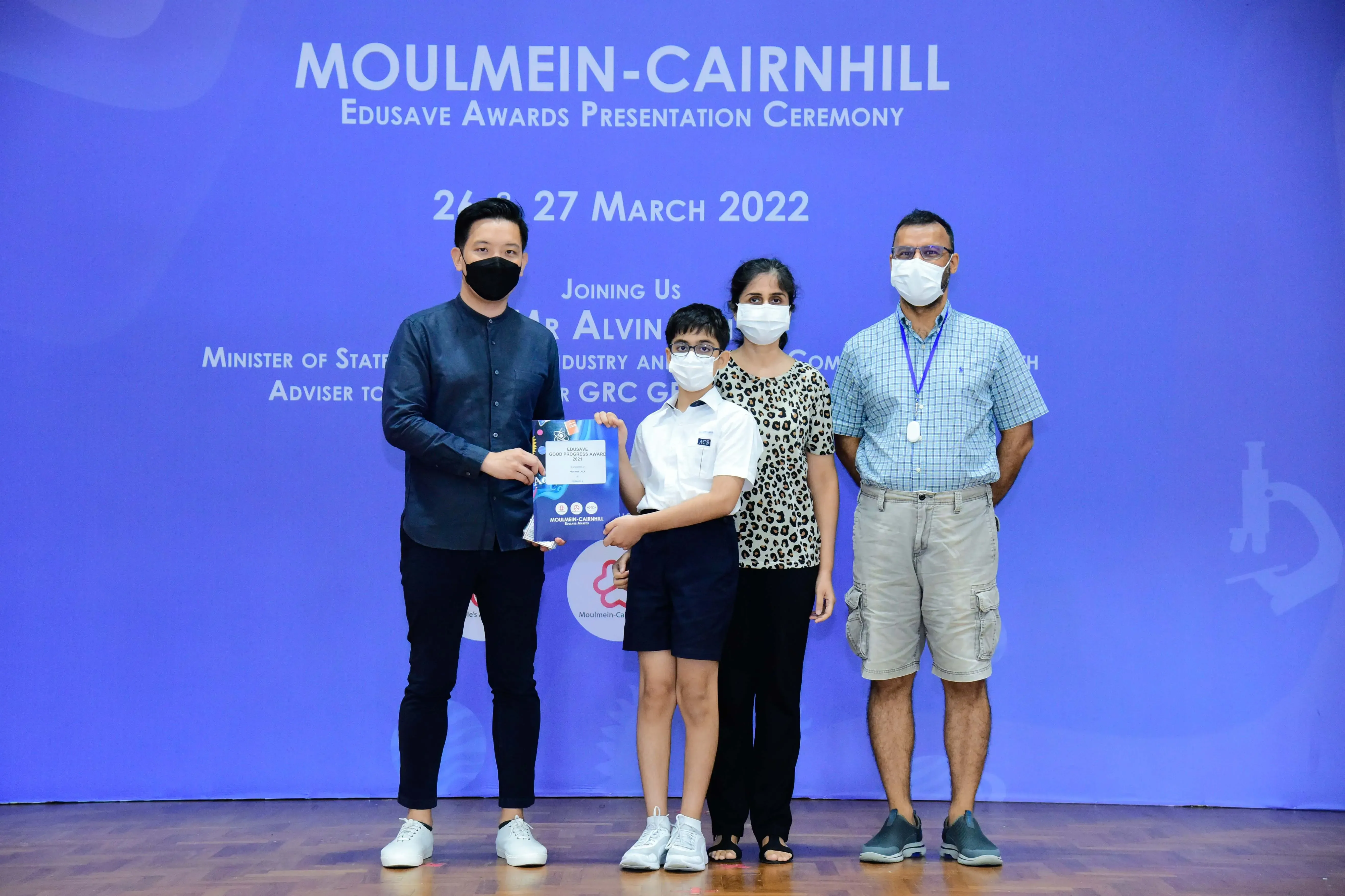 Four masked people pose at the Moulmein-Cairnhill Edusave Awards Ceremony, with a child holding a certificate.
