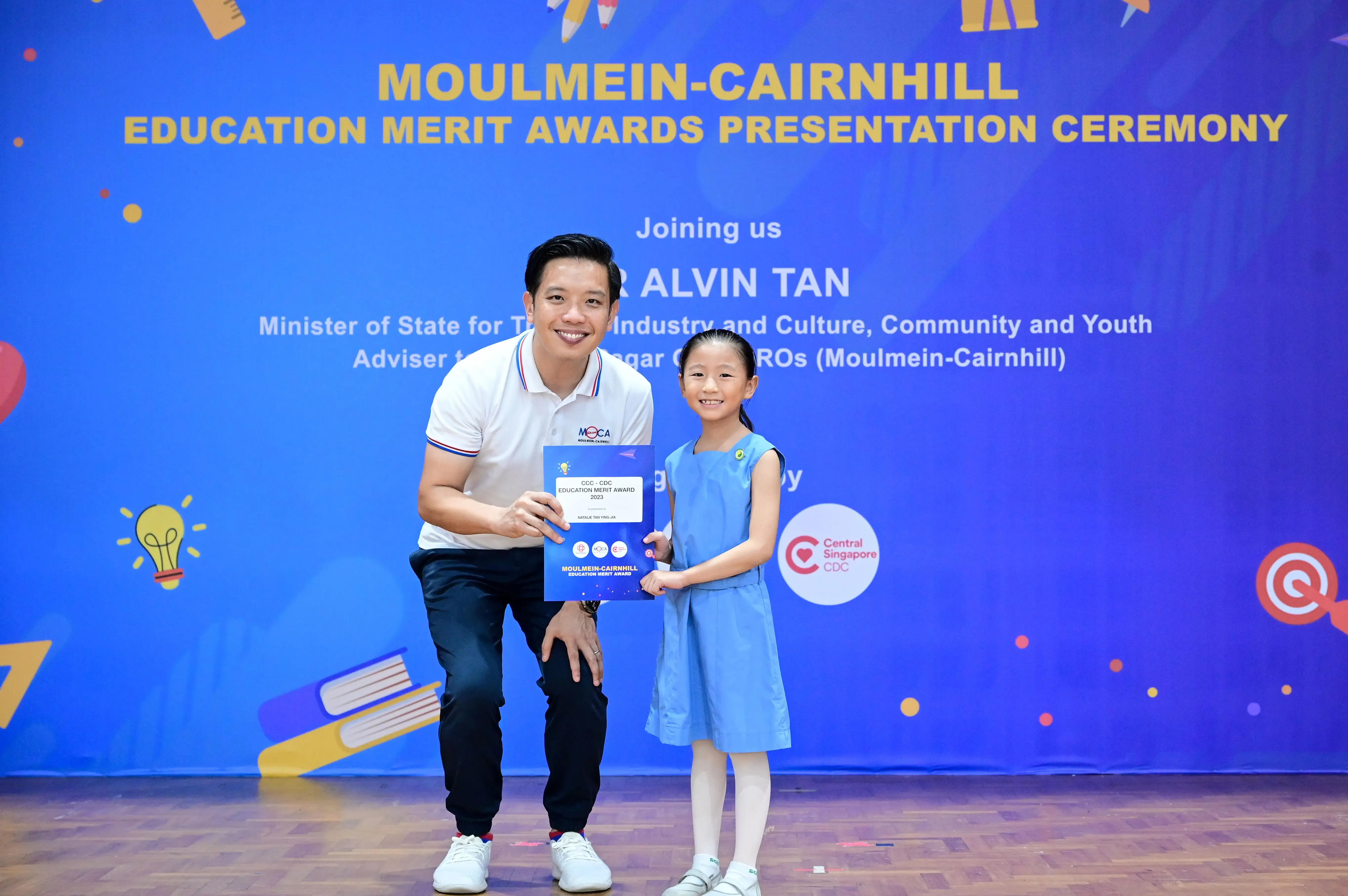 A man and a young girl hold a merit award certificate at the Moulmein-Cairnhill Education Merit Awards Ceremony.