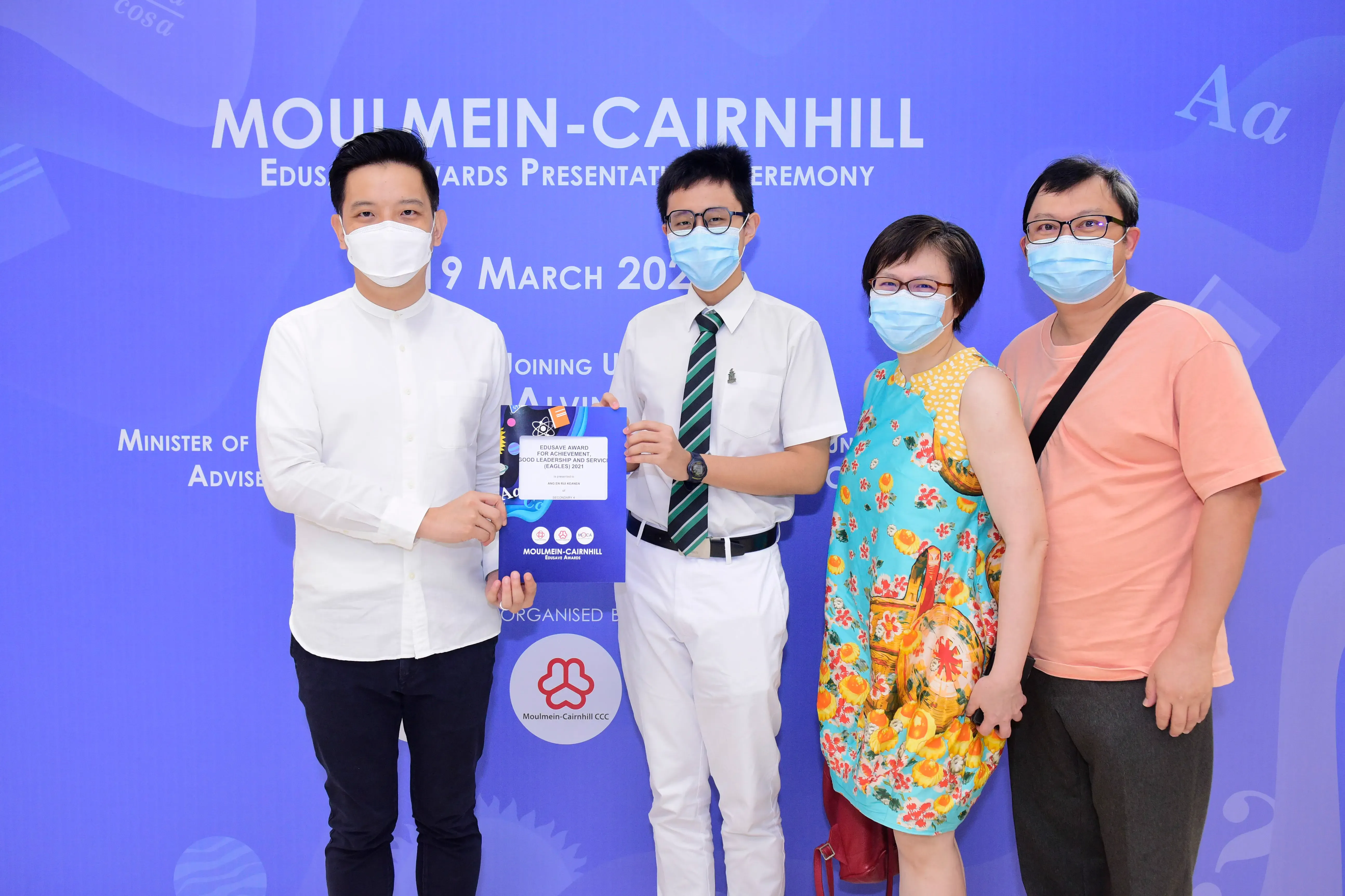 Four masked people stand before a Moulmein-Cairnhill Edusave Awards Presentation Ceremony banner, holding a certificate.