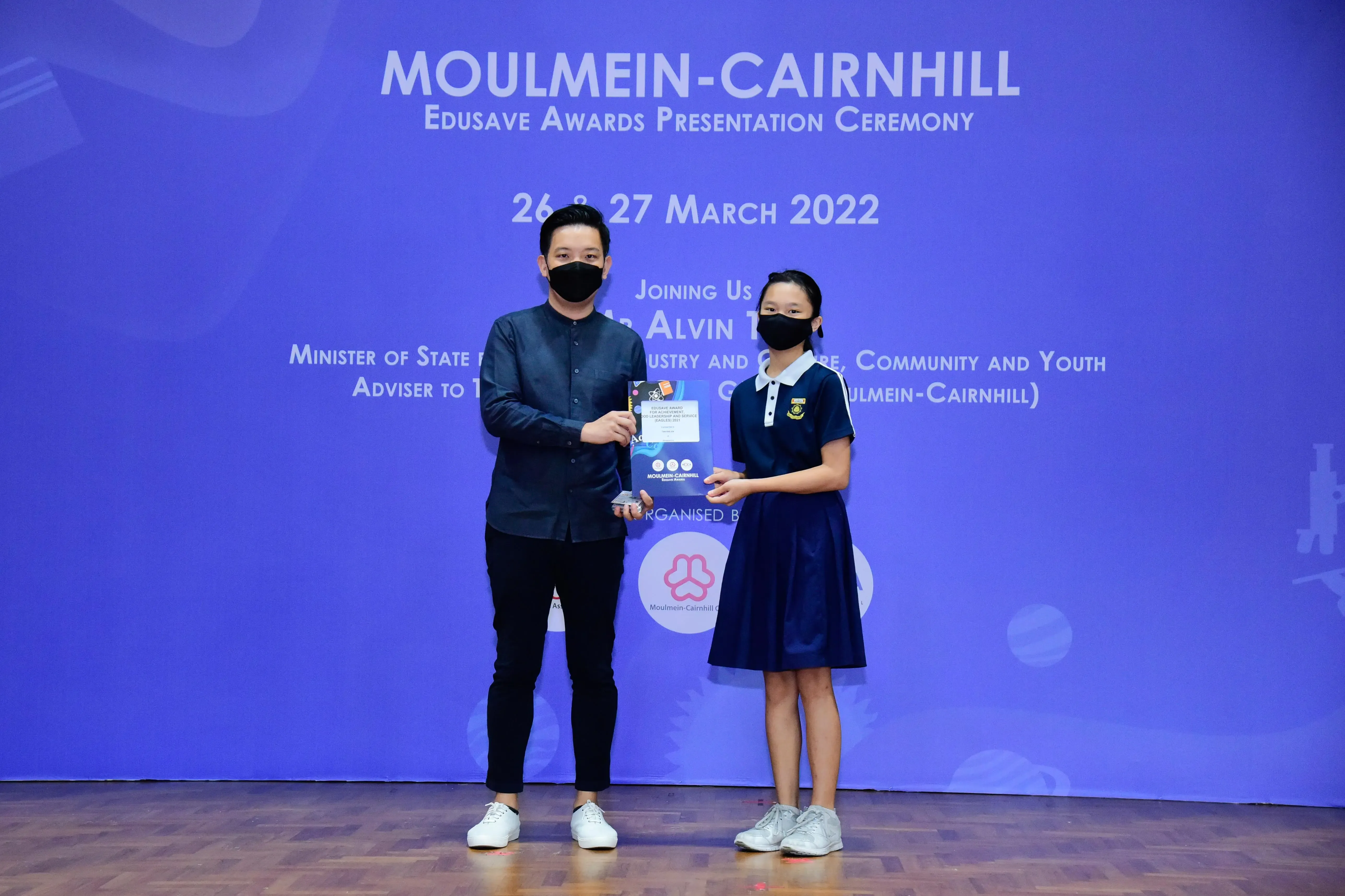 A masked adult and a school girl in uniform hold an award together at the Moulmein-Cairnhill Edusave Awards 2022.