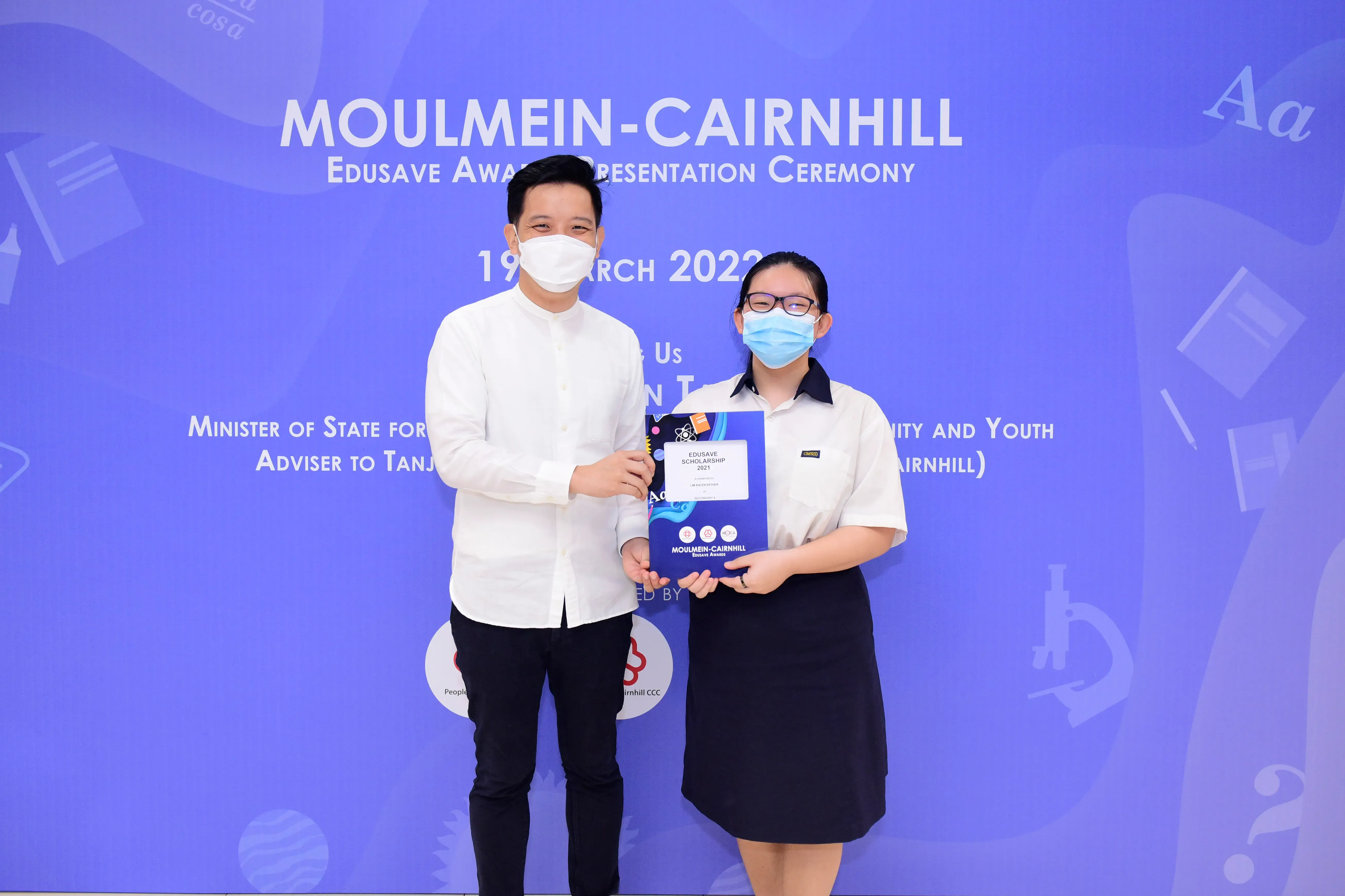 Two masked individuals hold an Edusave Scholarship certificate at the Moulmein-Cairnhill Award Ceremony, March 2022.