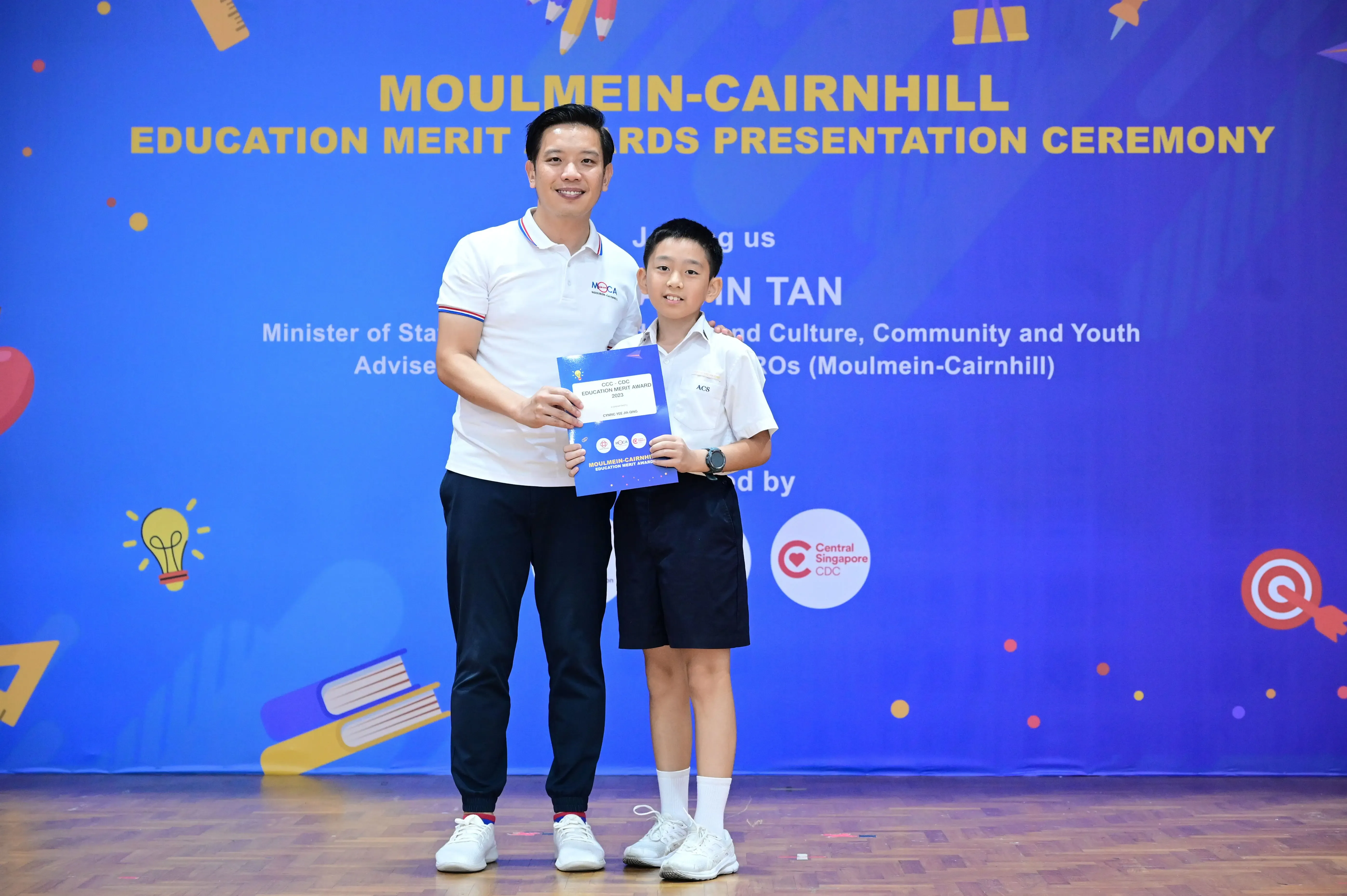 An adult and a young student hold a certificate at the Moulmein-Cairnhill Education Merit Awards Presentation Ceremony.