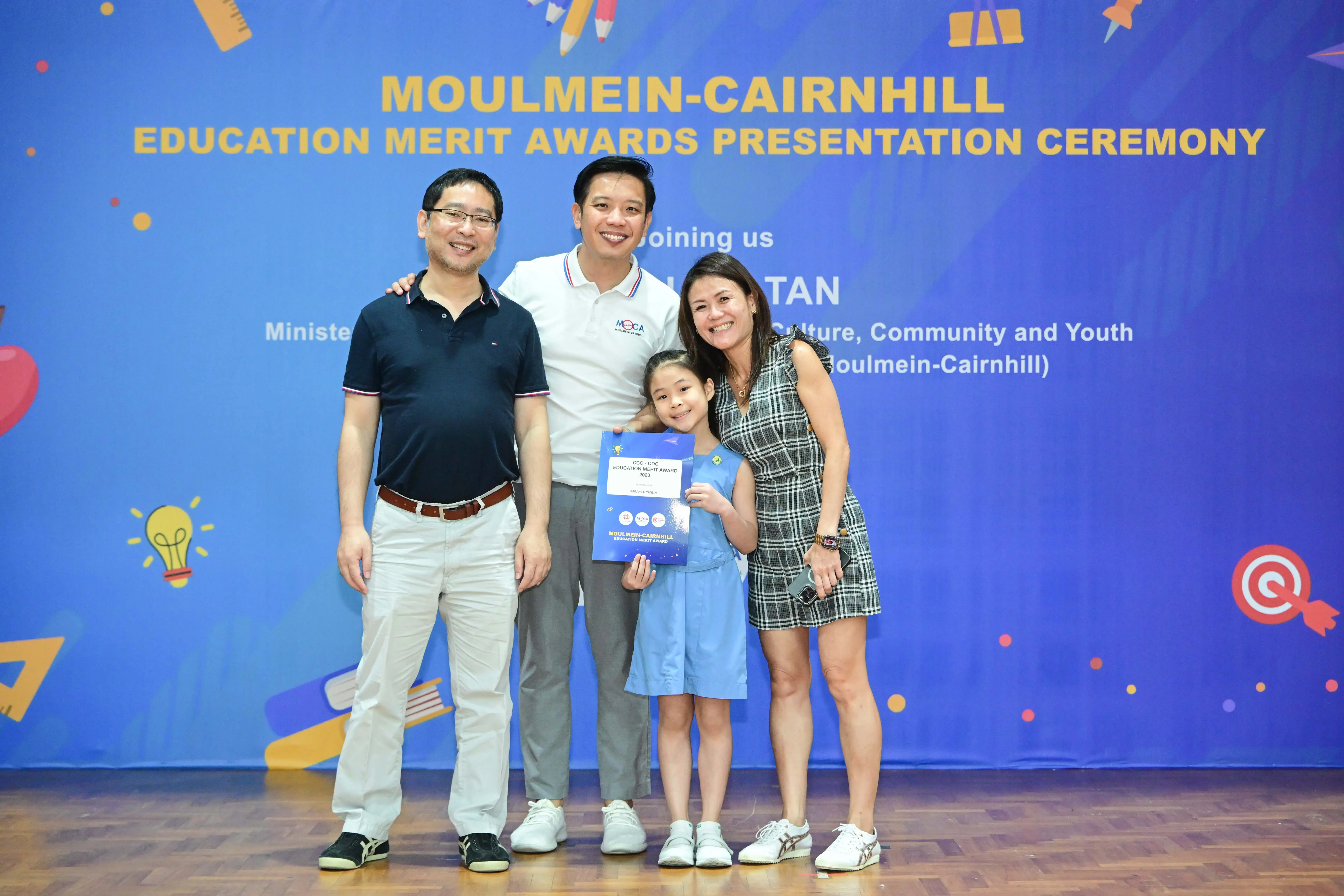 Four people pose smiling before a Moulmein-Cairnhill Education Merit Awards banner, with a young girl holding a blue certificate.