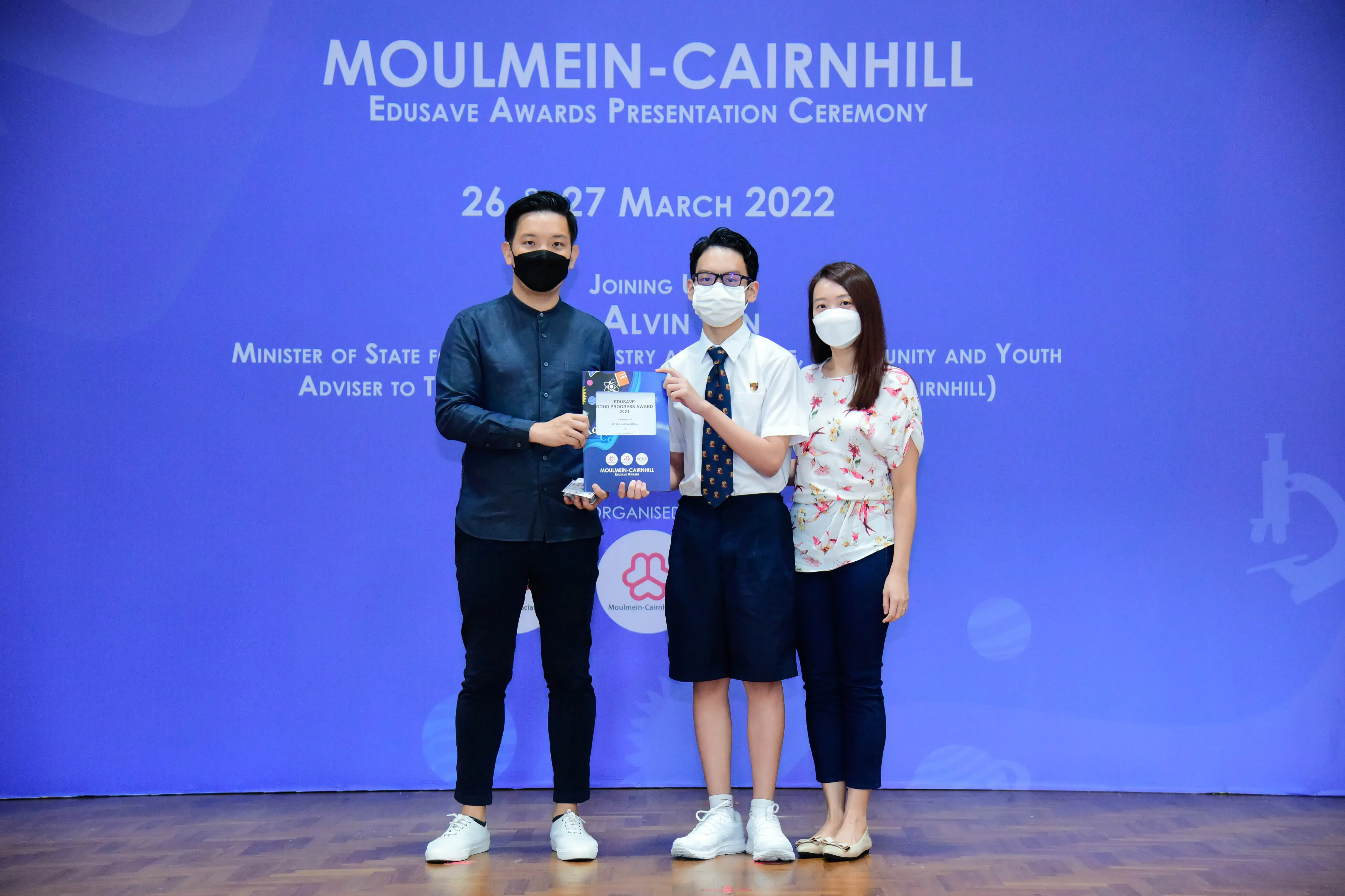 Three masked people pose on stage at the Moulmein-Cairnhill Edusave Awards Presentation Ceremony, March 2022.