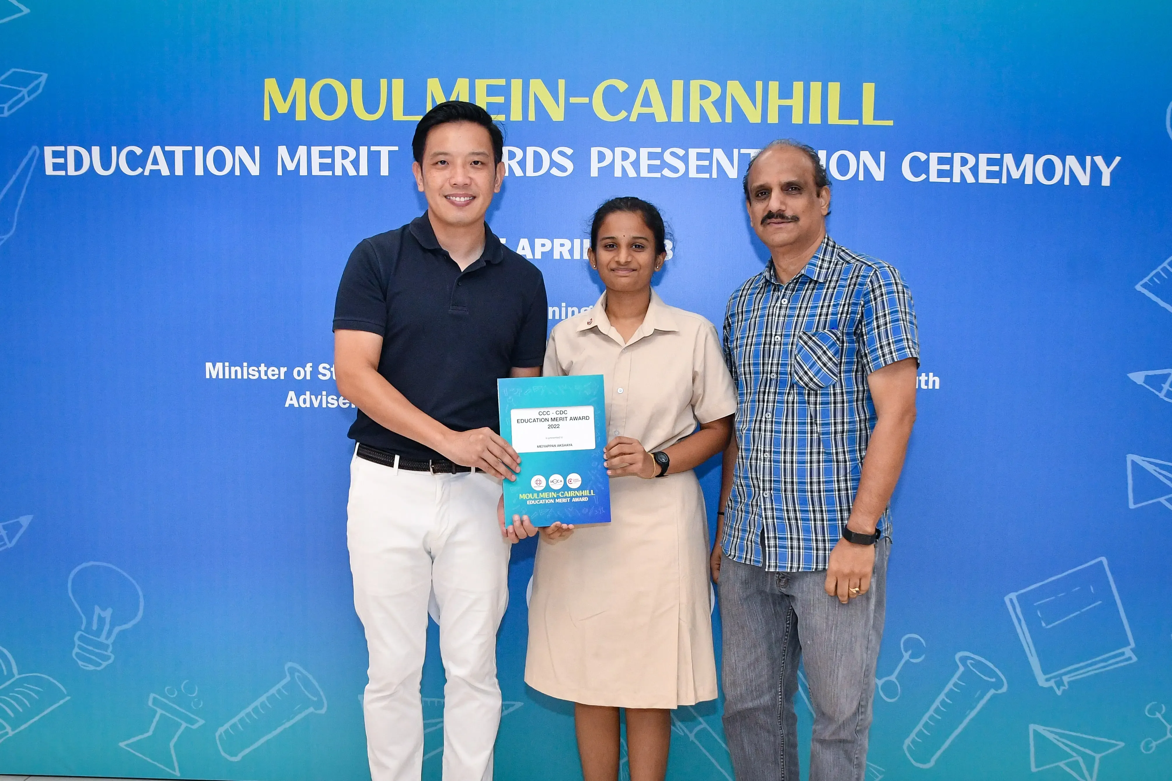 Three people pose at the Moulmein-Cairnhill Education Merit Awards Presentation Ceremony, holding a blue award folder.