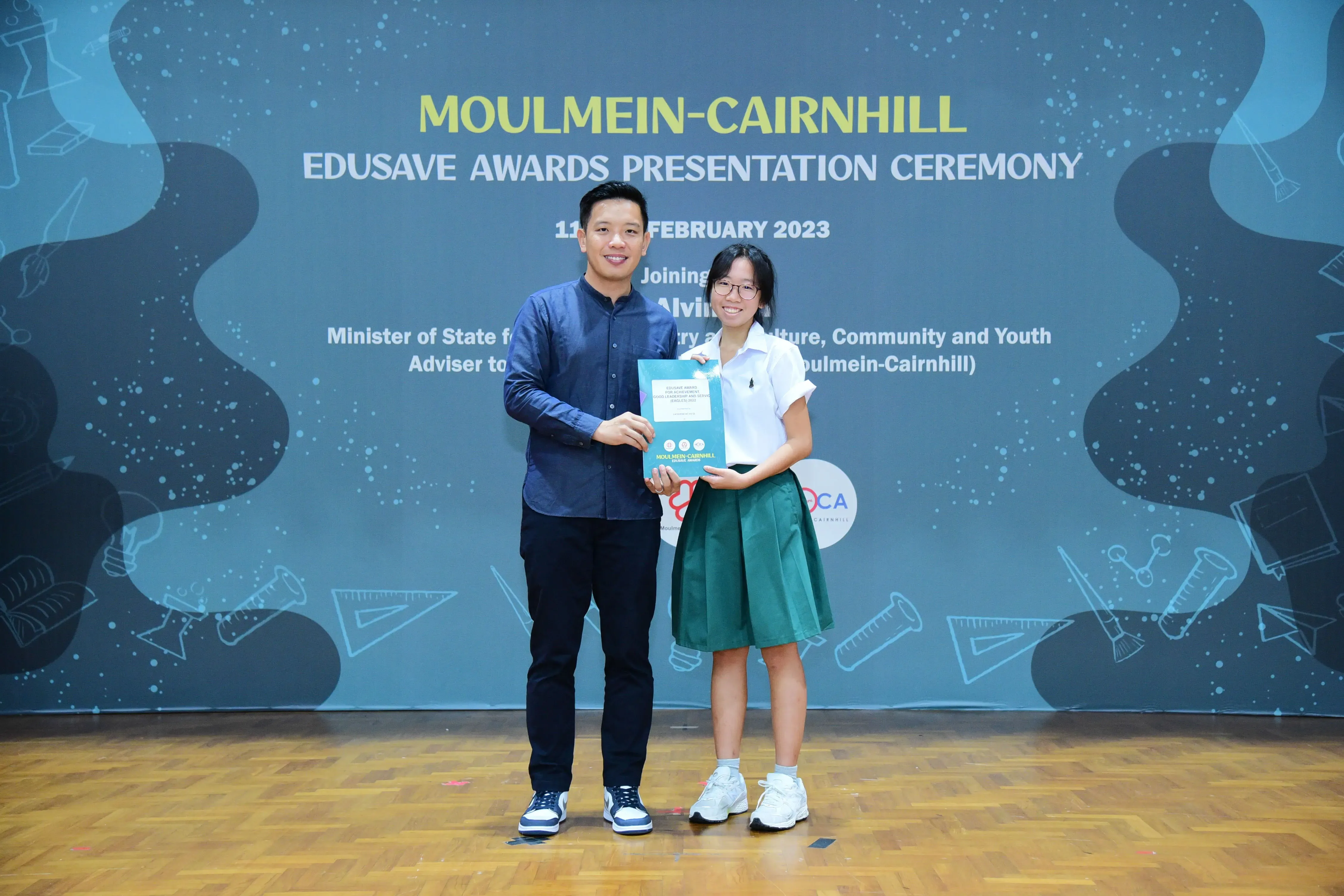 Two people stand holding a certificate at the Moulmein-Cairnhill Edusave Awards Presentation Ceremony, February 2023.