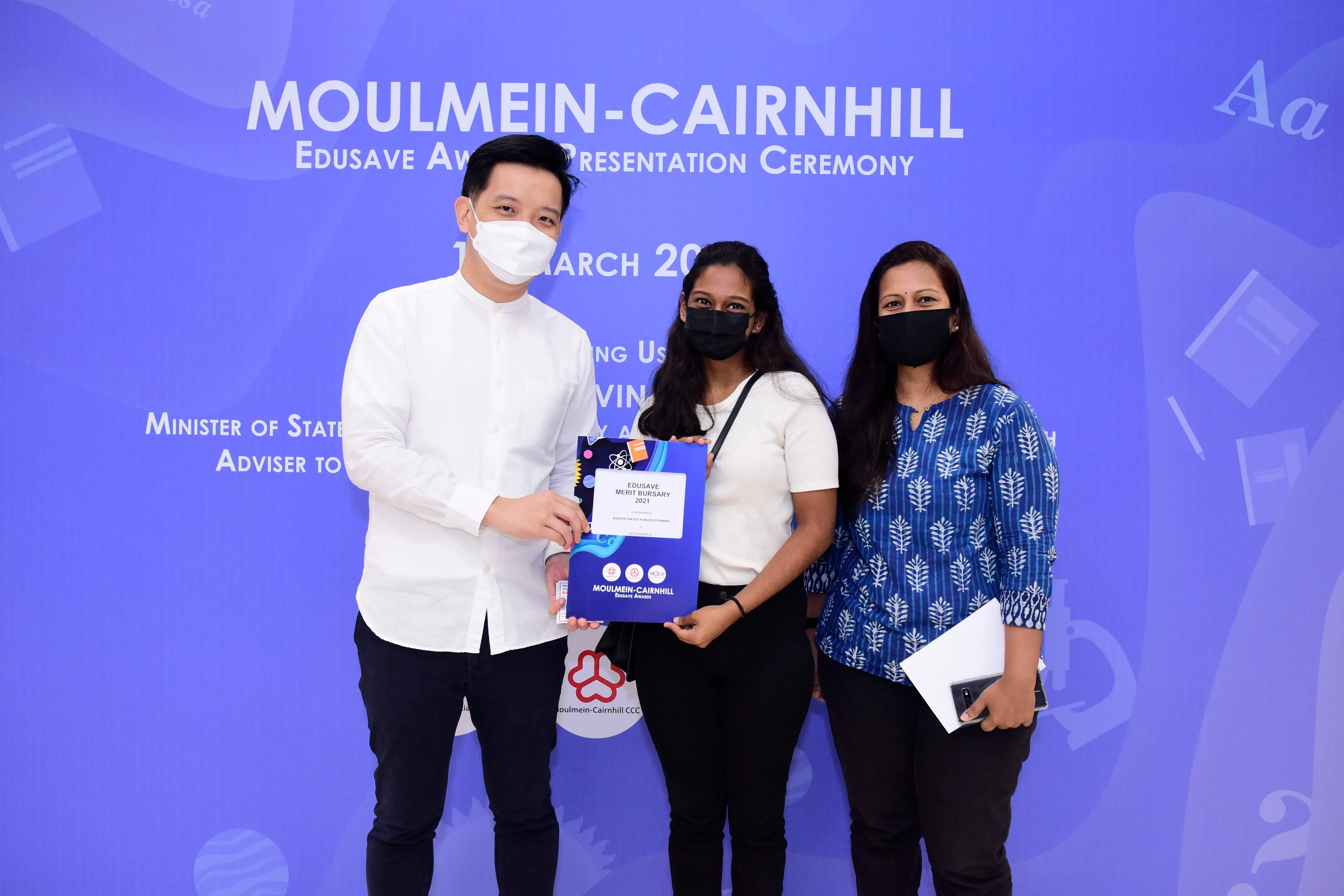Three masked people pose holding an Edusave Merit Bursary certificate at the Moulmein-Cairnhill Award Ceremony.