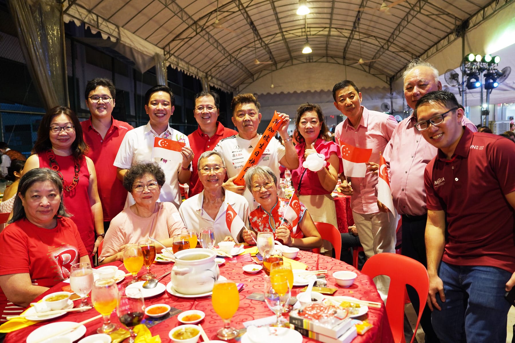Group of people in red clothing gathered at a dinner table, holding Singapore flags, celebrating National Day.