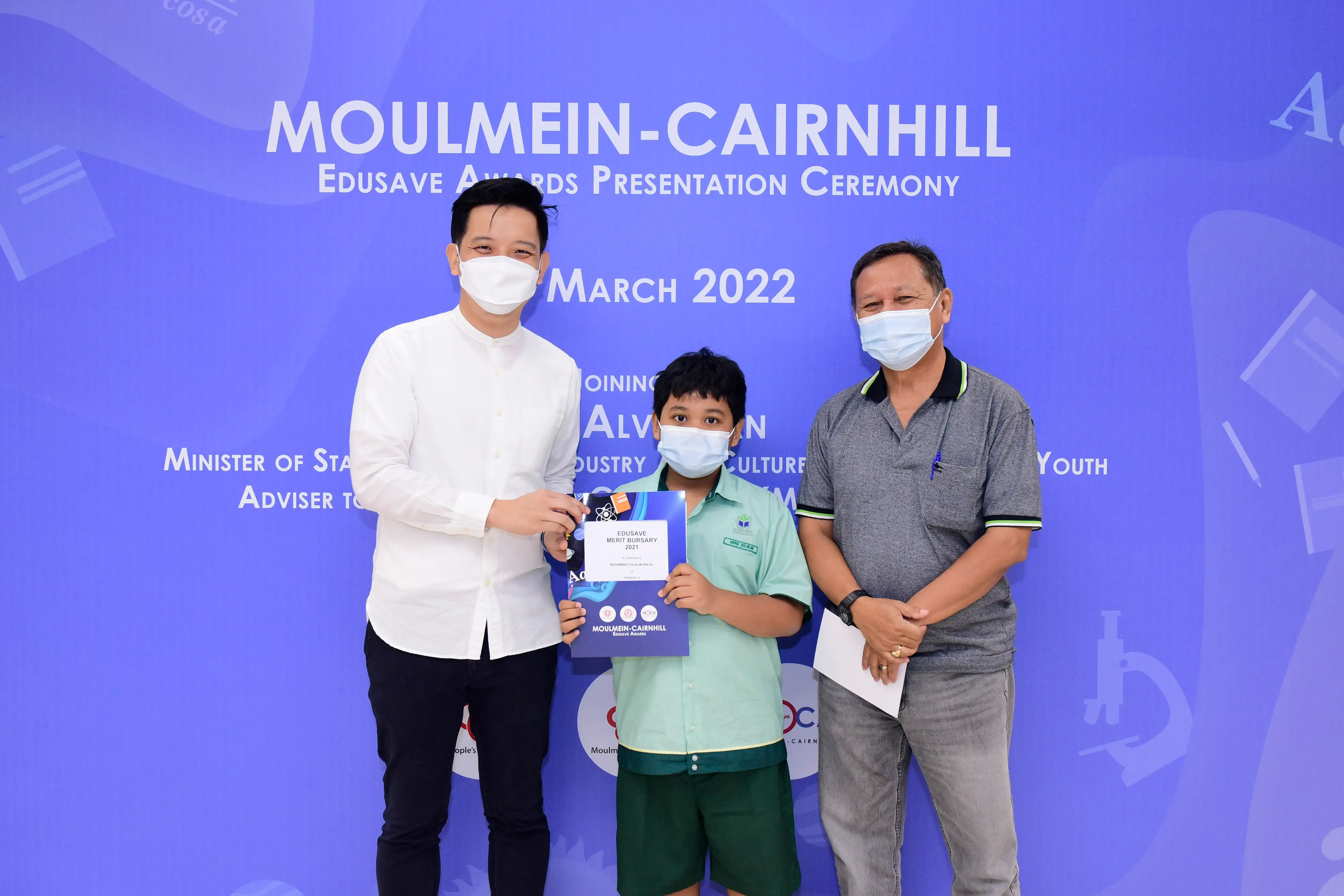 Two masked adults and a masked child holding an Edusave Merit Bursary certificate at a March 2022 awards ceremony.