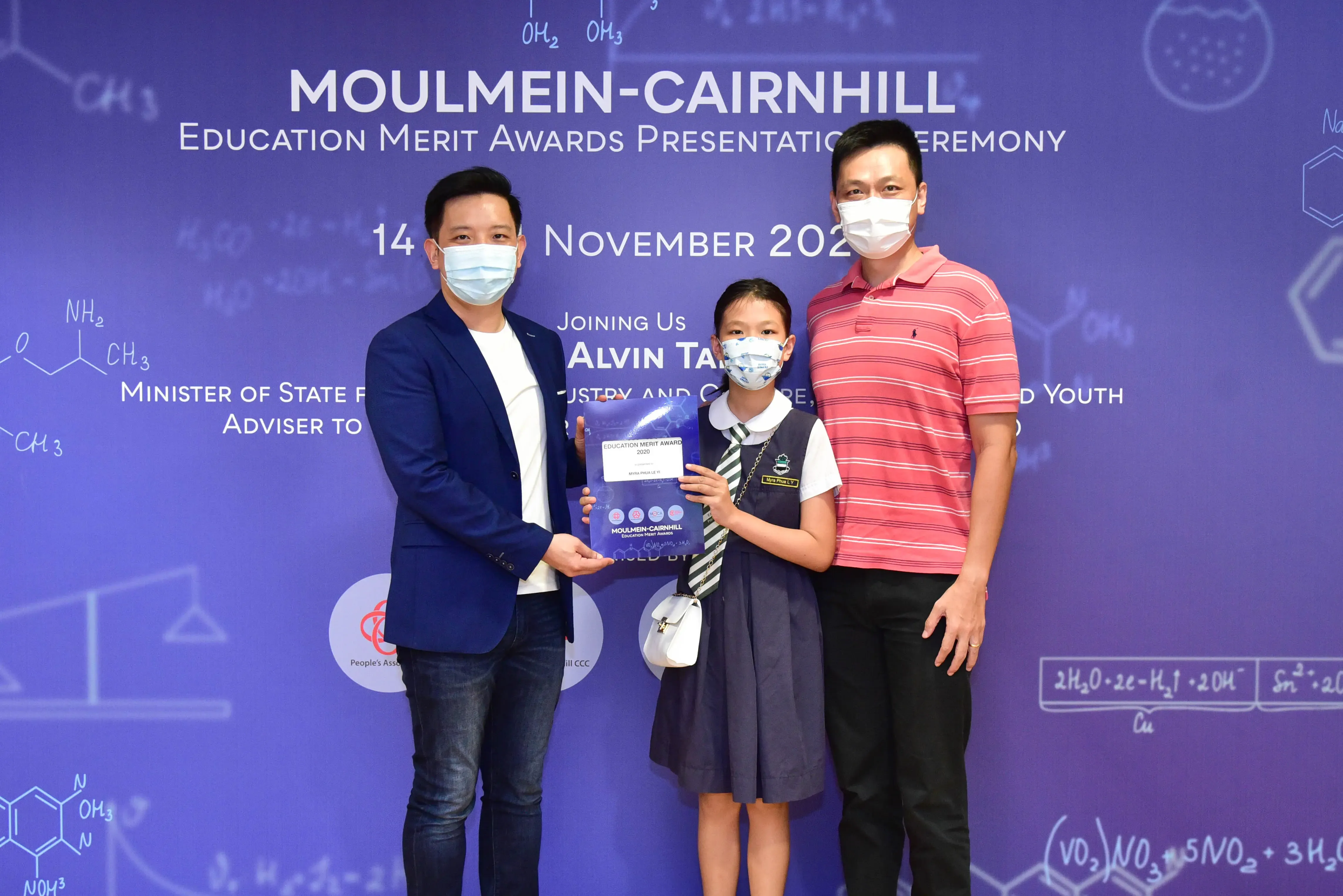 Two masked men and a masked schoolgirl hold a certificate at the Moulmein-Cairnhill Education Merit Awards ceremony.