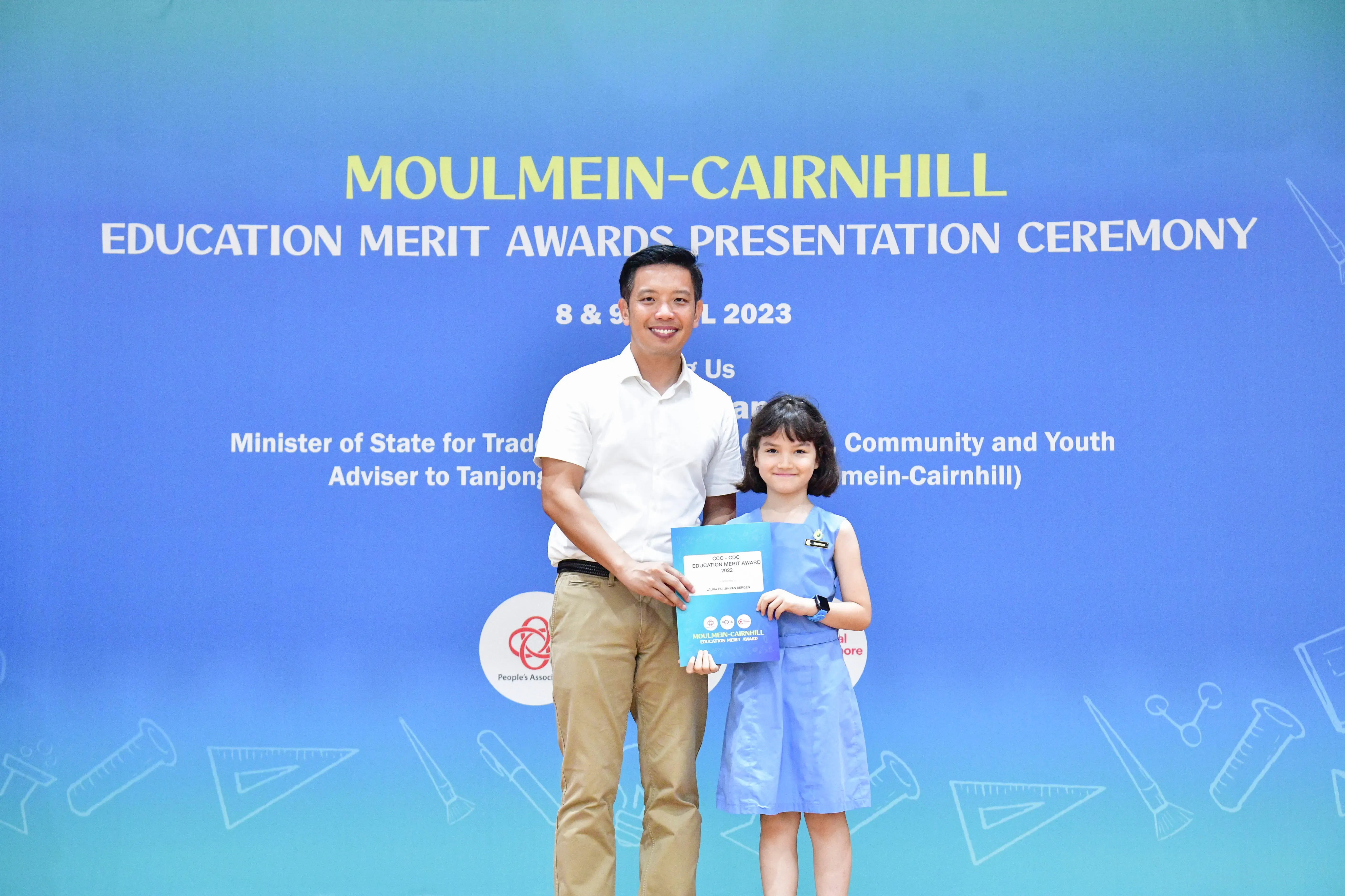 A man and a young girl hold an award certificate in front of a Moulmein-Cairnhill Education Merit Awards banner.