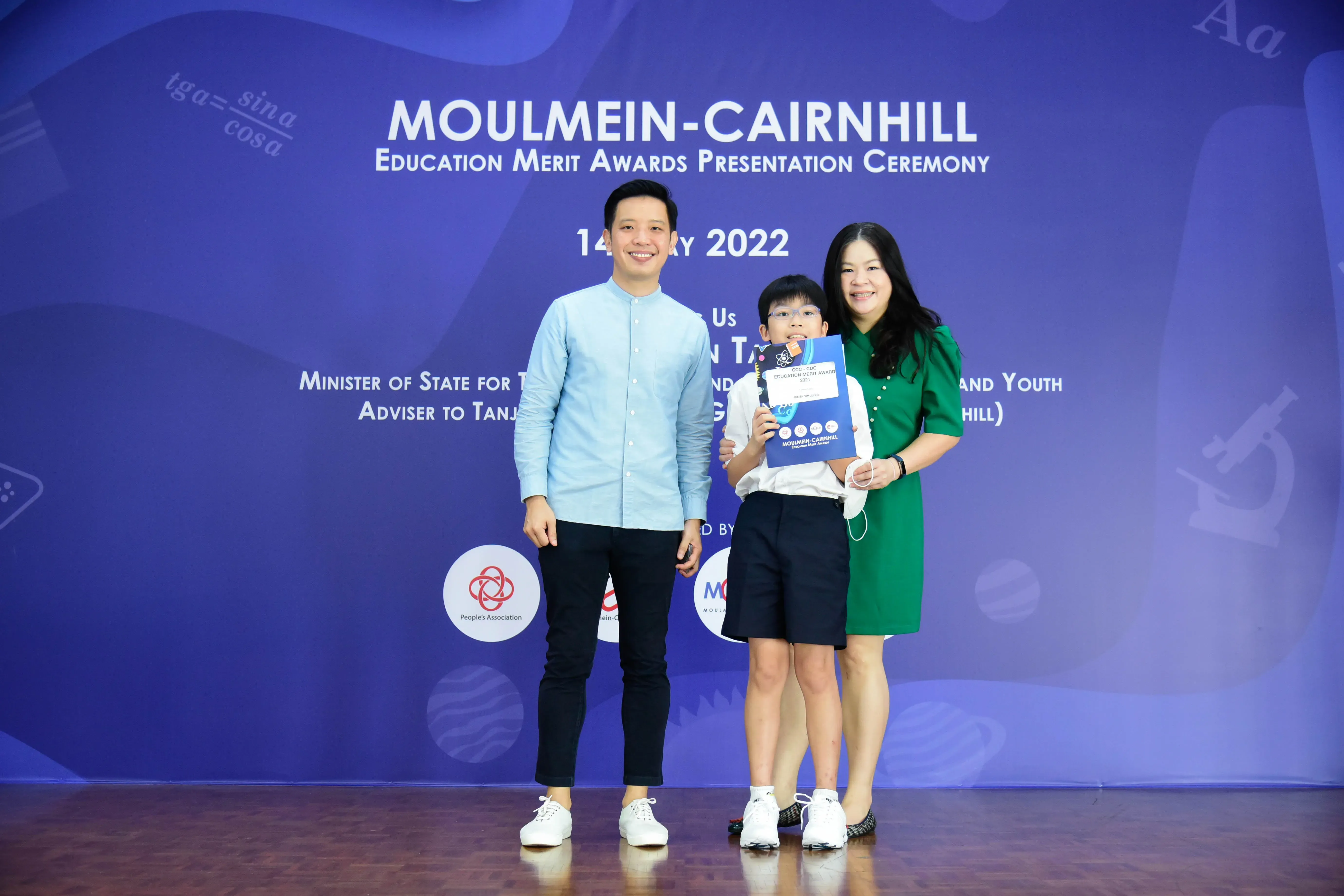 Two adults and a child pose before a Moulmein-Cairnhill Education Merit Awards 2022 ceremony backdrop.