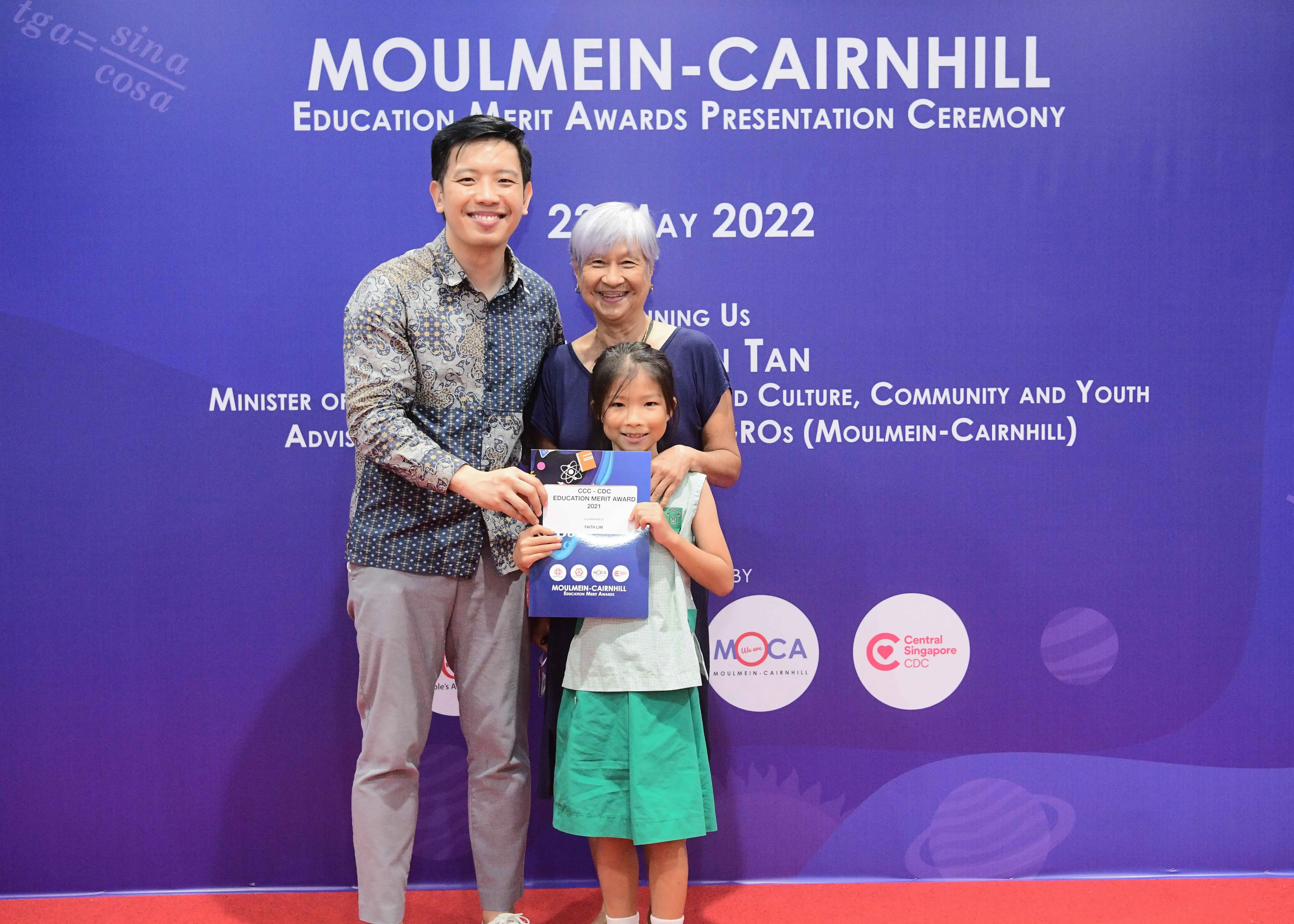 Three people pose smiling at the Moulmein-Cairnhill Education Merit Awards Ceremony in May 2022, child holding an award.