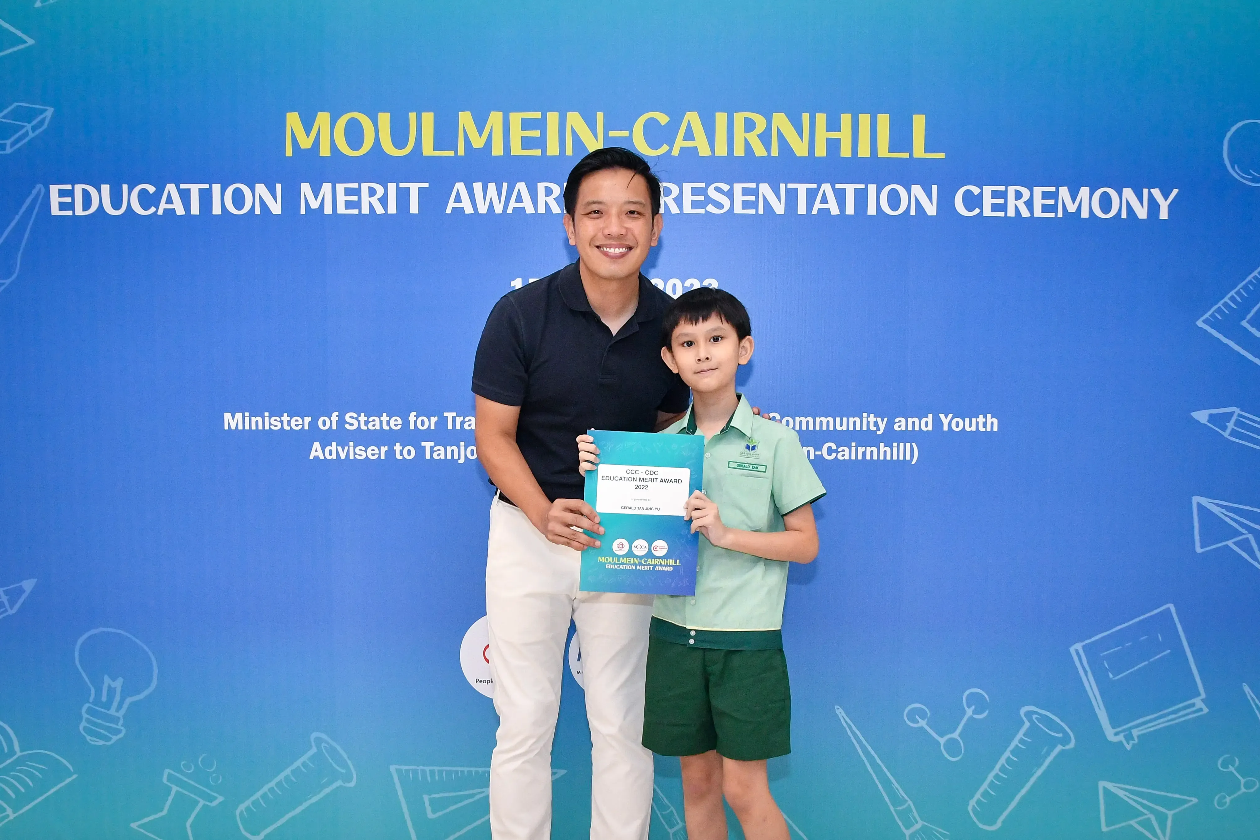 A man and a young boy in school uniform hold an award certificate at the Moulmein-Cairnhill Education Merit Award ceremony.