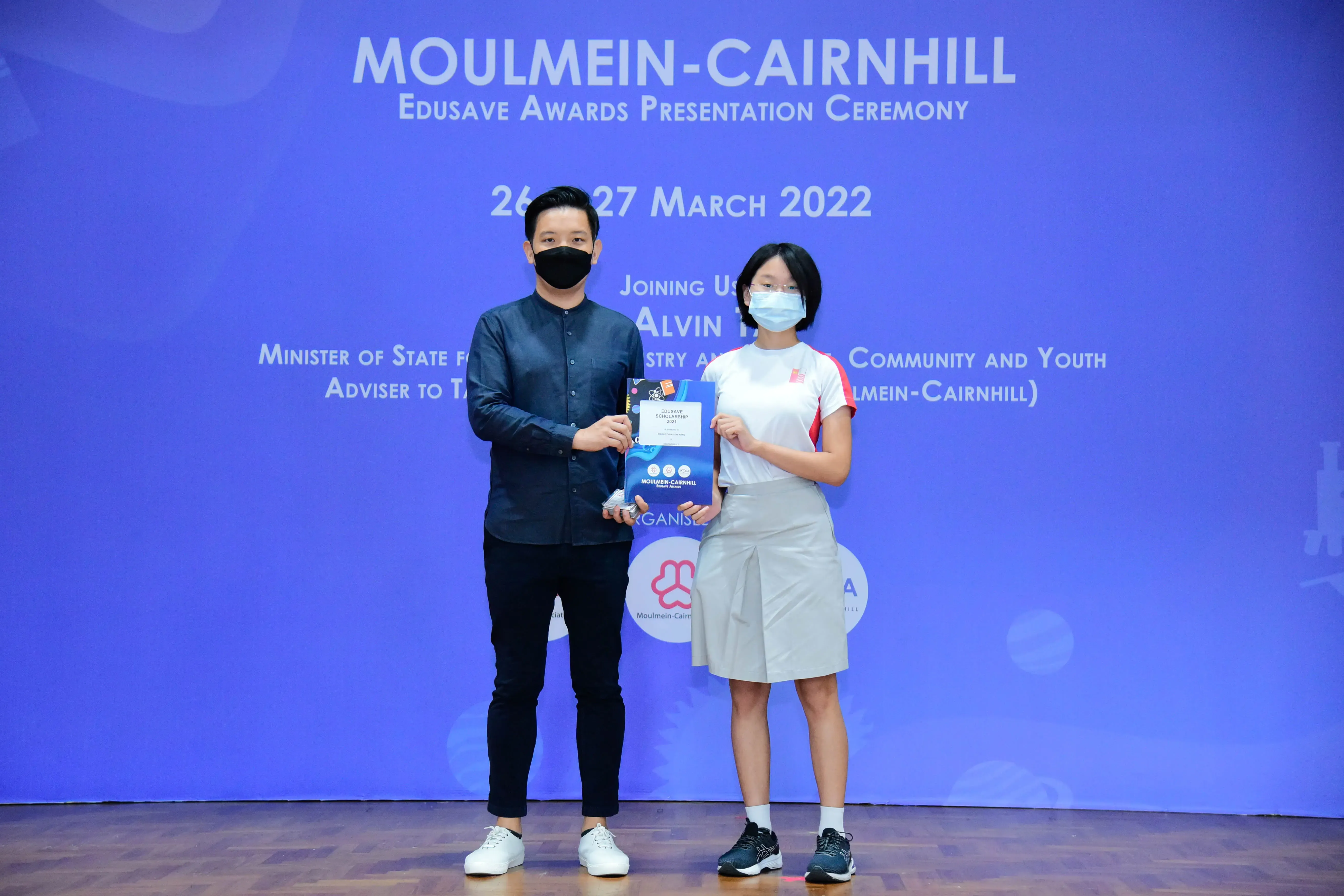 Two masked people hold a certificate on stage at the Moulmein-Cairnhill Edusave Awards Ceremony, March 2022.