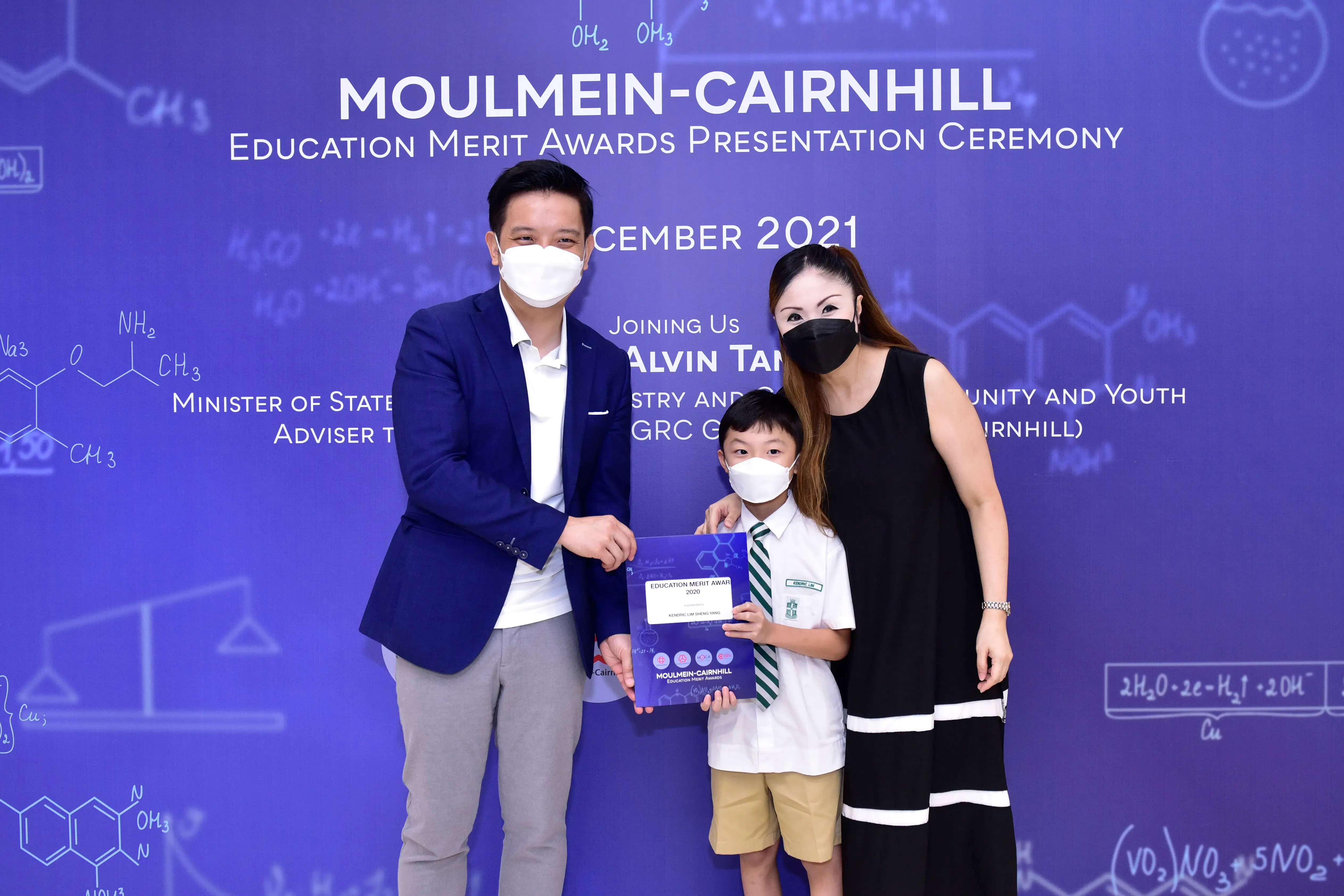Three masked people pose at the Moulmein-Cairnhill Education Merit Awards Ceremony 2021, with a child holding a certificate.