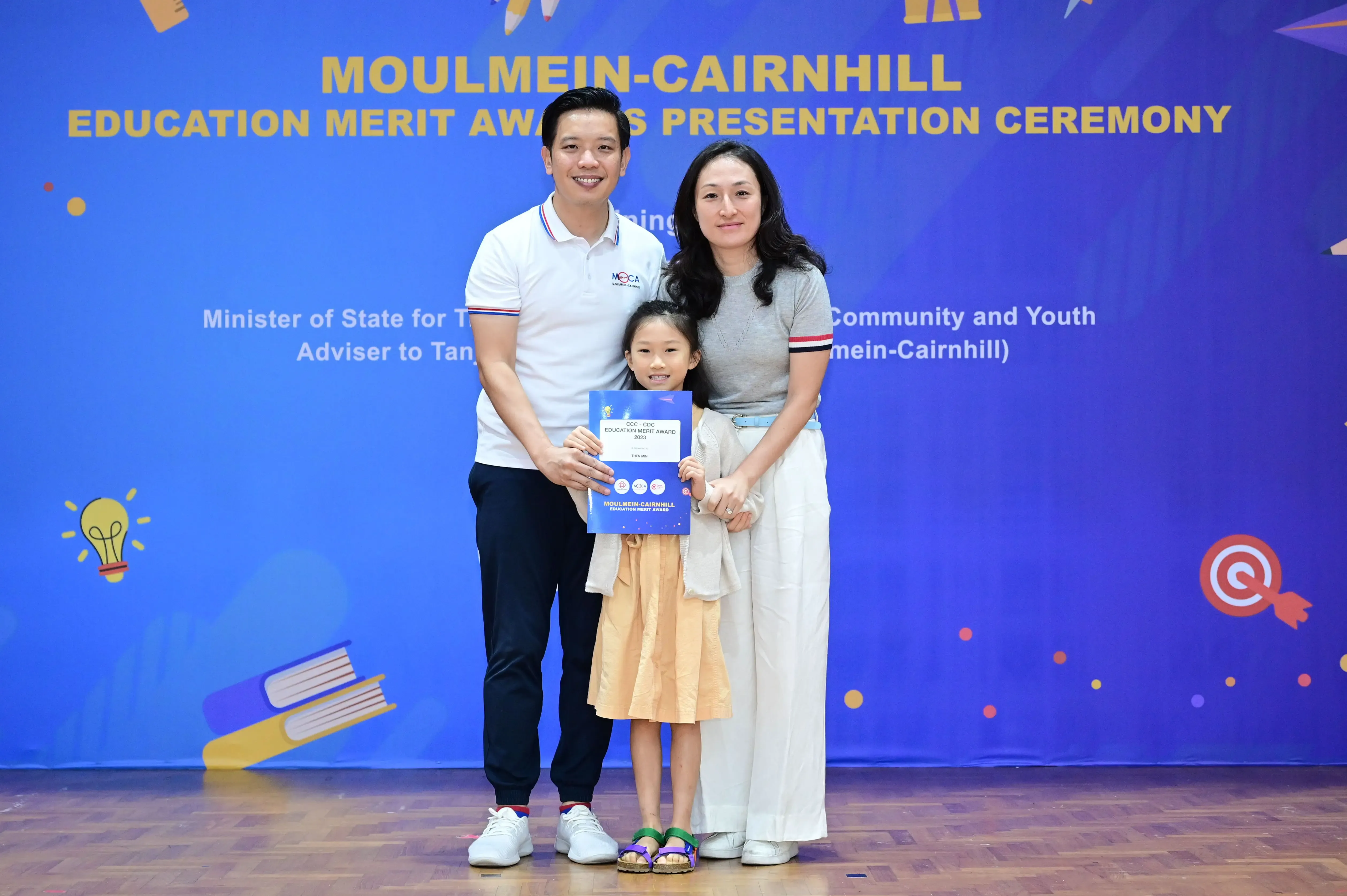 Two adults and a young girl pose with a certificate at the Moulmein-Cairnhill Education Merit Awards ceremony.