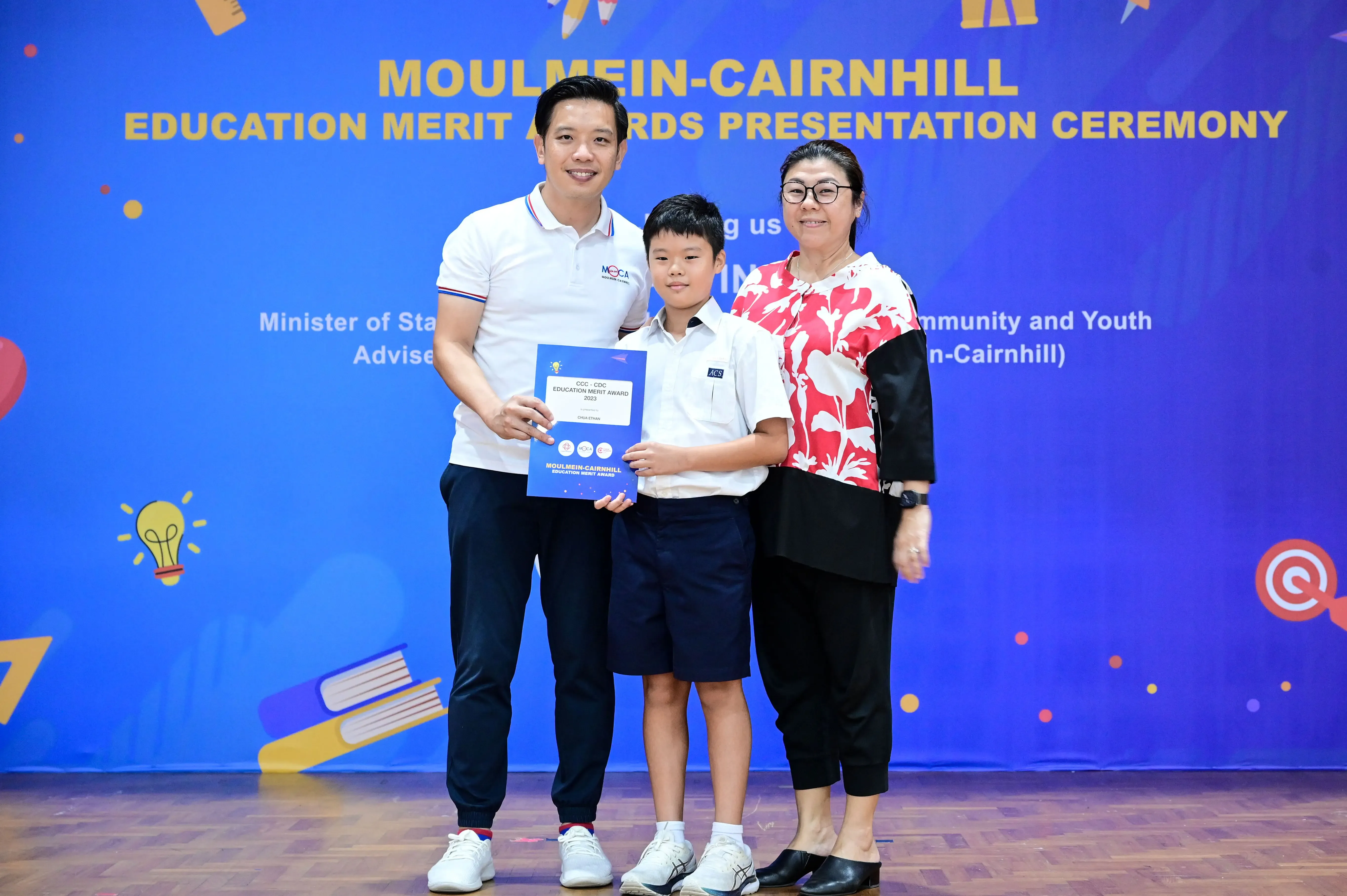 Two adults and a young boy holding an award certificate at the Moulmein-Cairnhill Education Merit Awards ceremony.