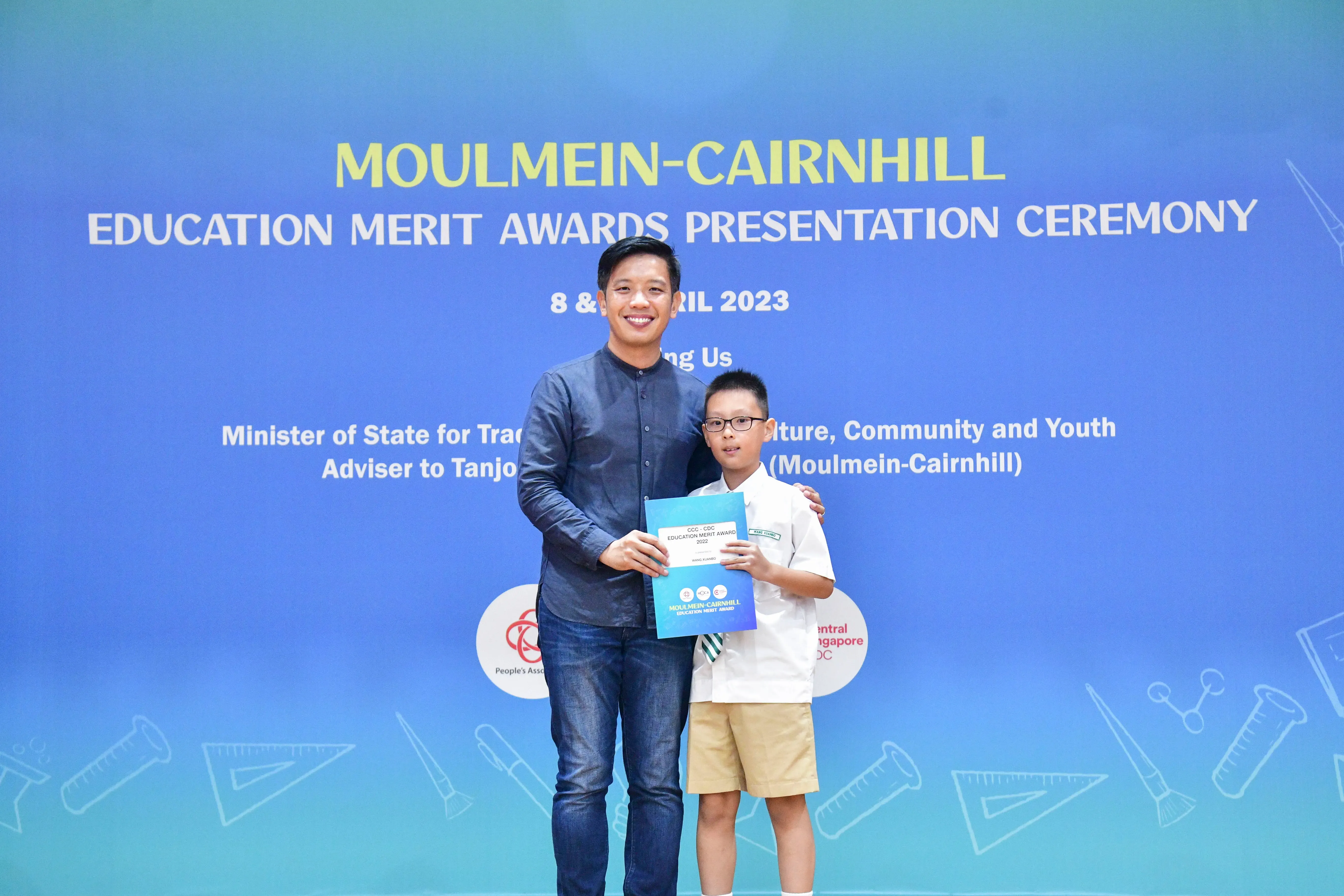 A man and a young boy hold a certificate in front of a Moulmein-Cairnhill Education Merit Awards banner.