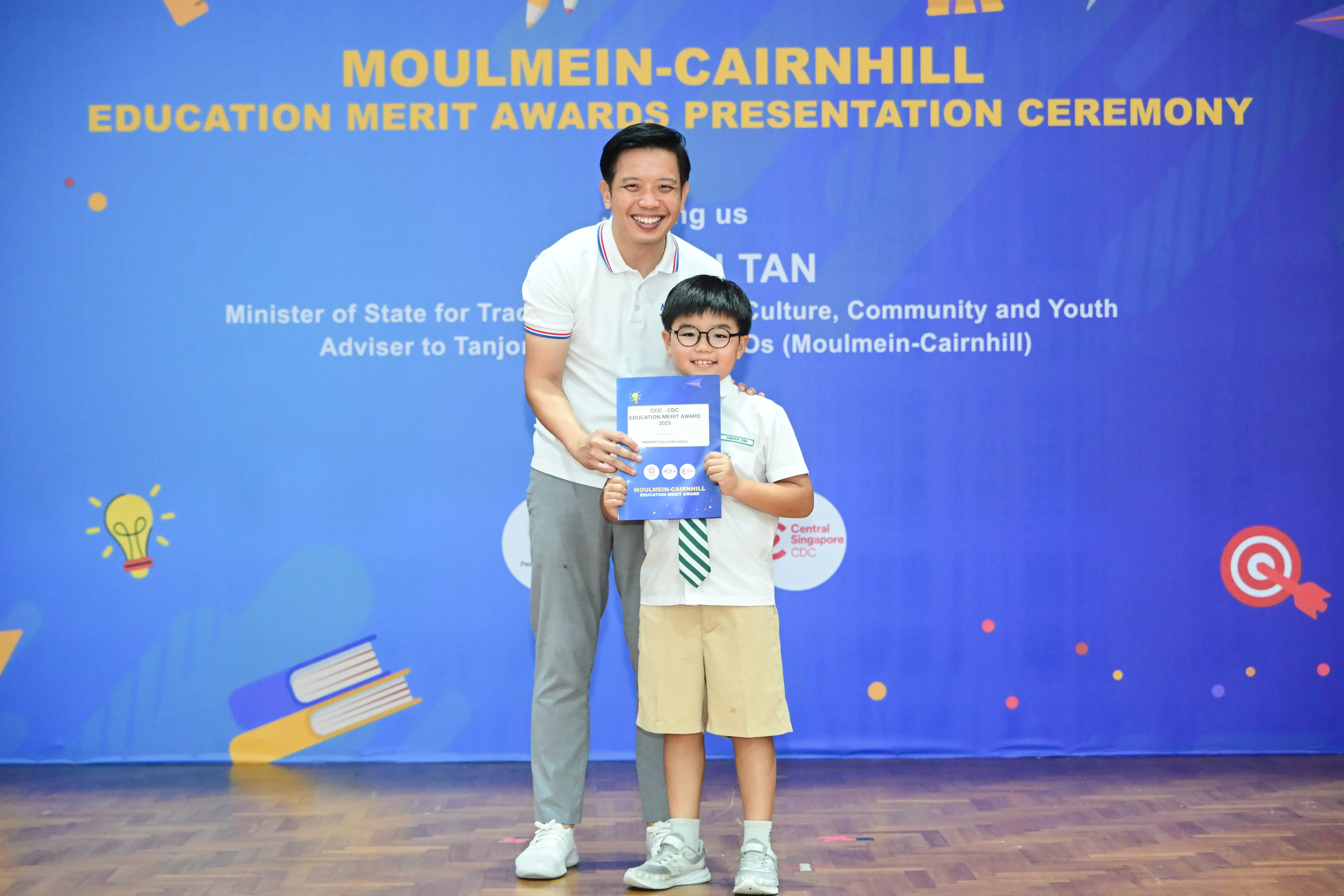 A man and a young boy holding a blue certificate at the Moulmein-Cairnhill Education Merit Awards Presentation Ceremony.