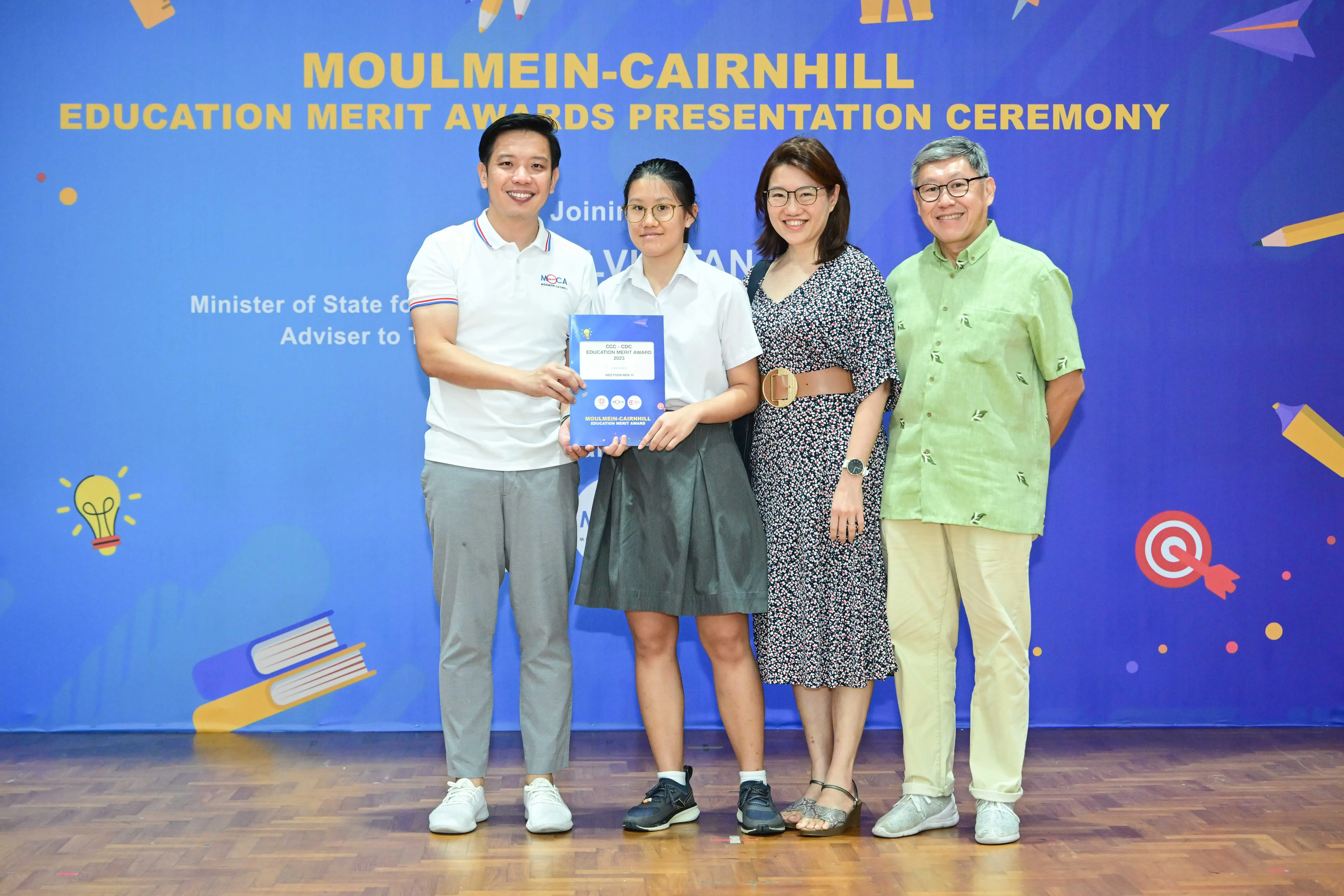 Four people pose smiling at the Moulmein-Cairnhill Education Merit Awards Presentation Ceremony, one holding a certificate.