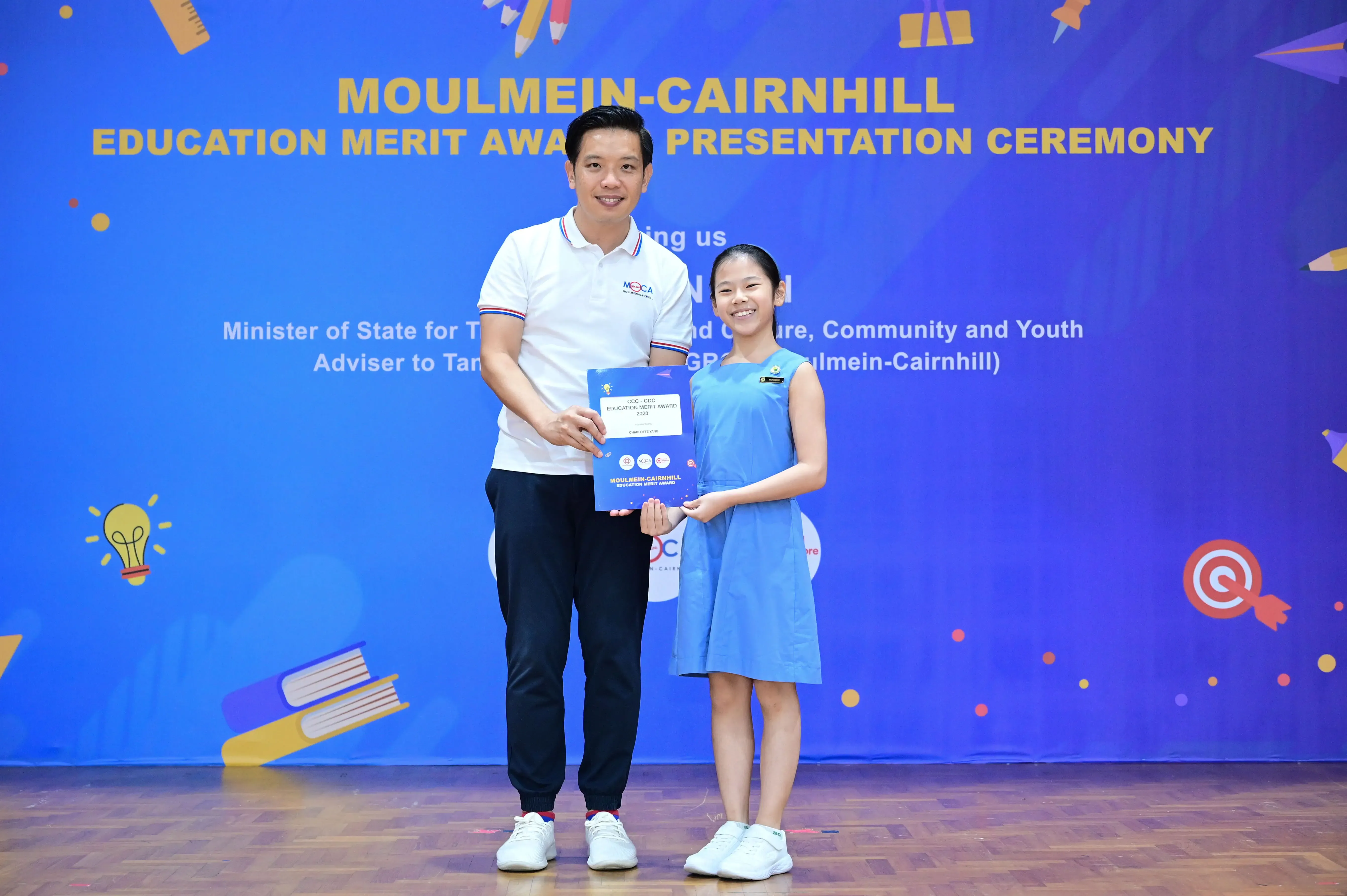 A man and a young girl hold a certificate together at the Moulmein-Cairnhill Education Merit Award ceremony.