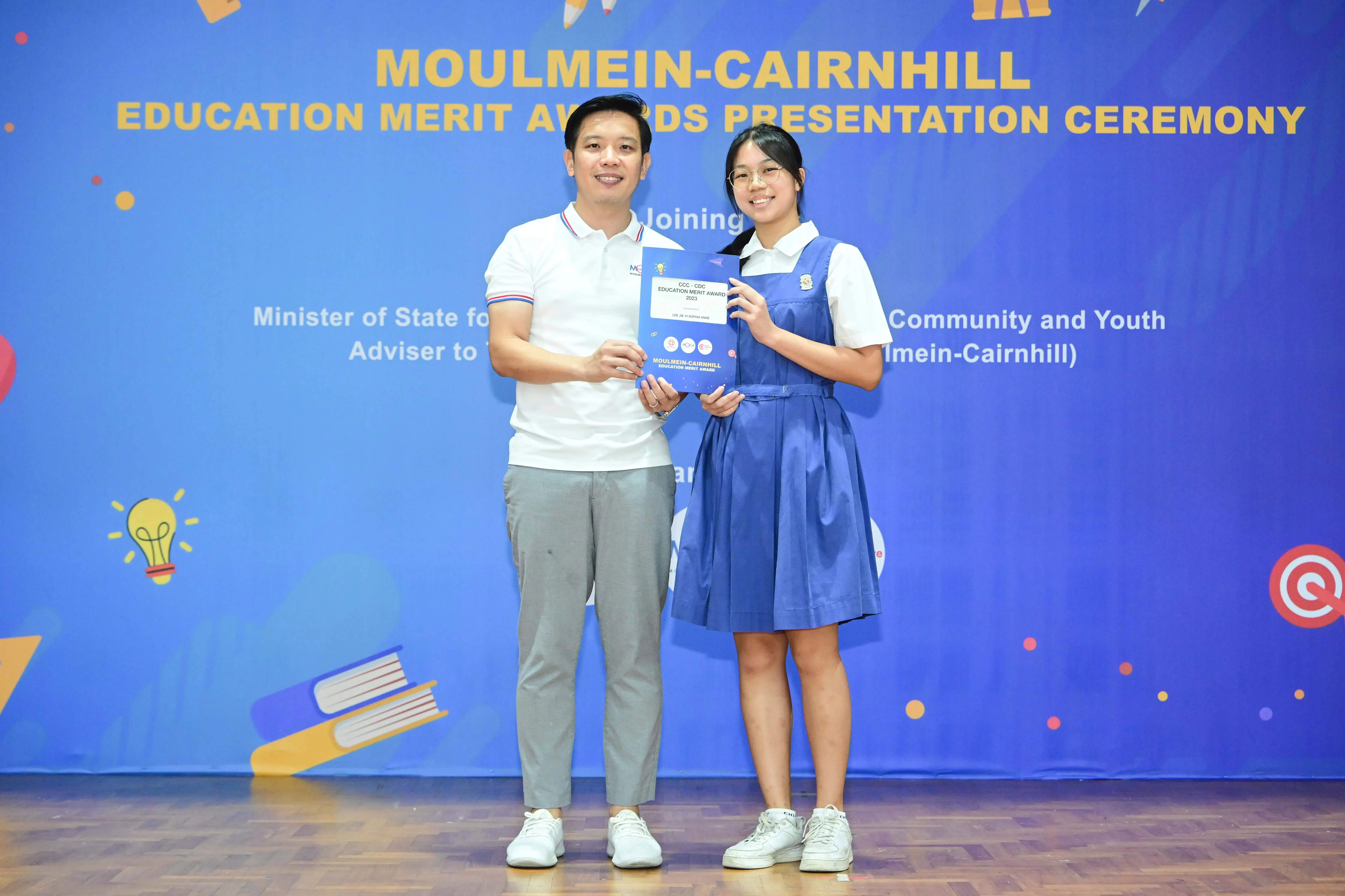 Two people hold a certificate in front of a Moulmein-Cairnhill Education Merit Awards Presentation Ceremony banner.