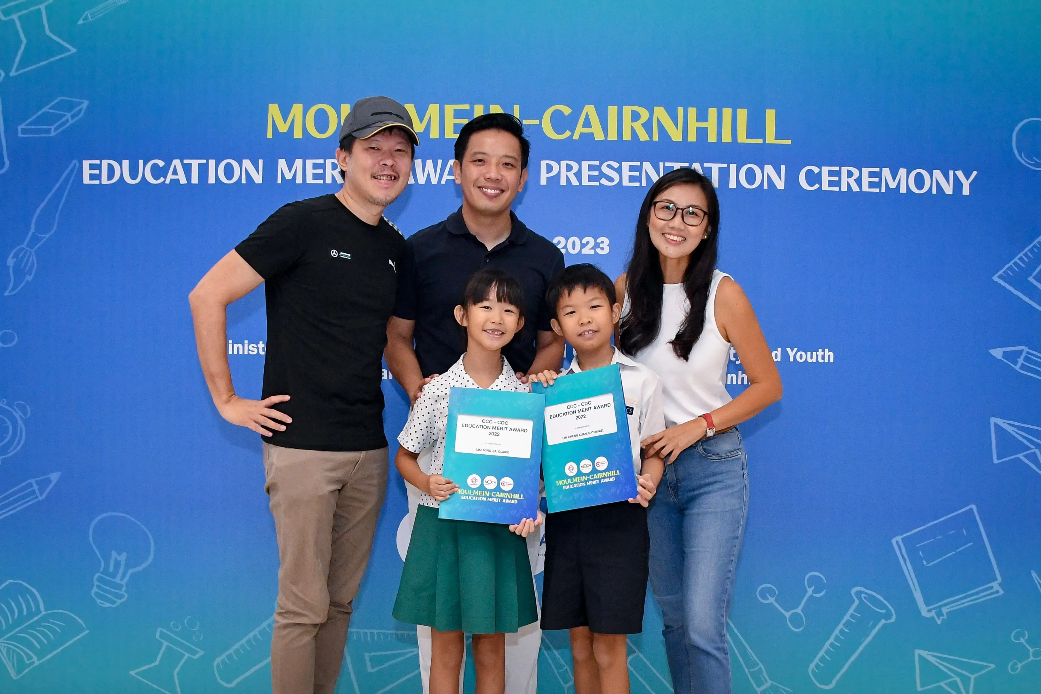 Two adults and two children pose at the 2023 Moulmein-Cairnhill Education Merit Award ceremony, kids holding certificates.