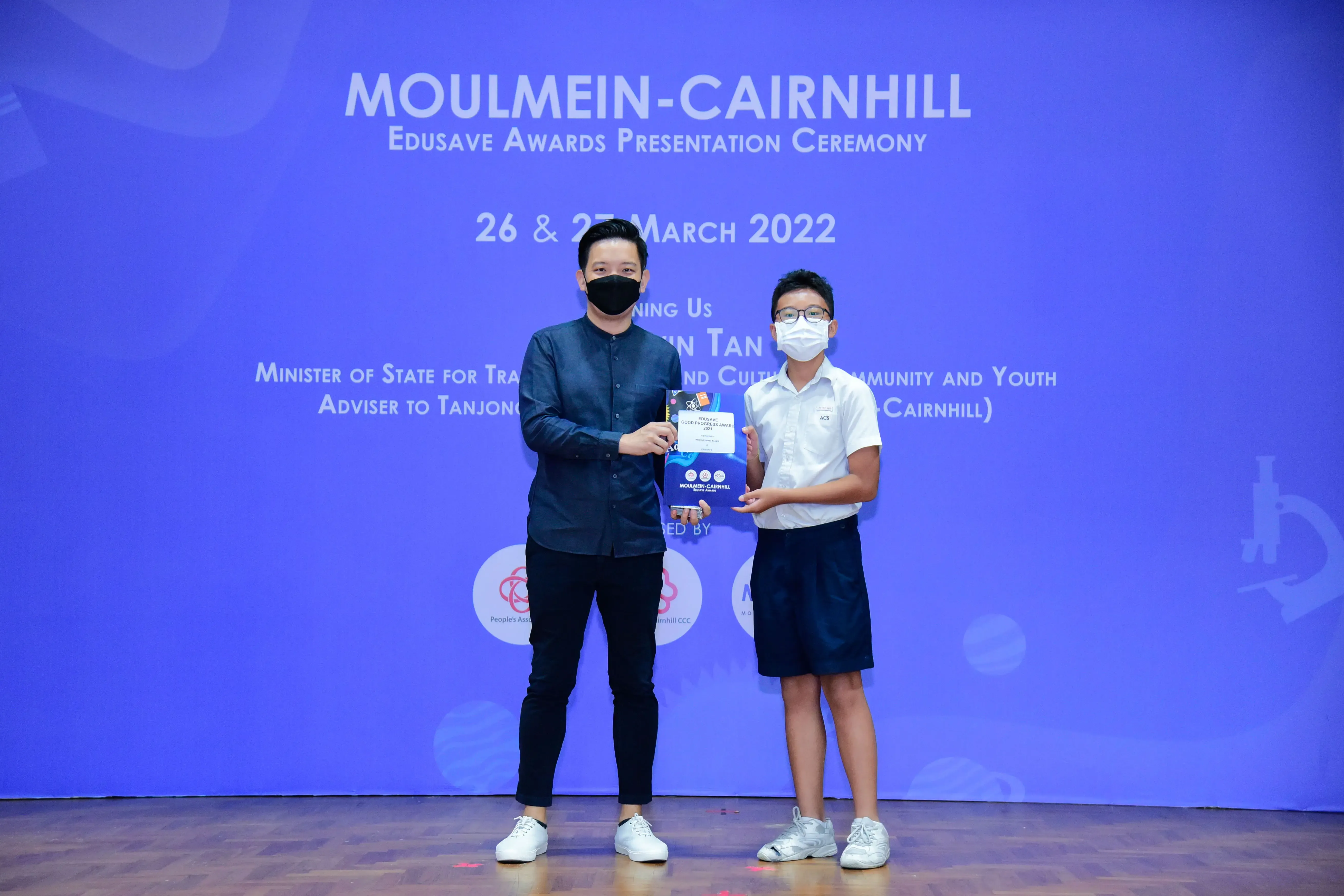 An adult and a young student in masks hold a certificate at the Moulmein-Cairnhill Edusave Awards Ceremony 2022.
