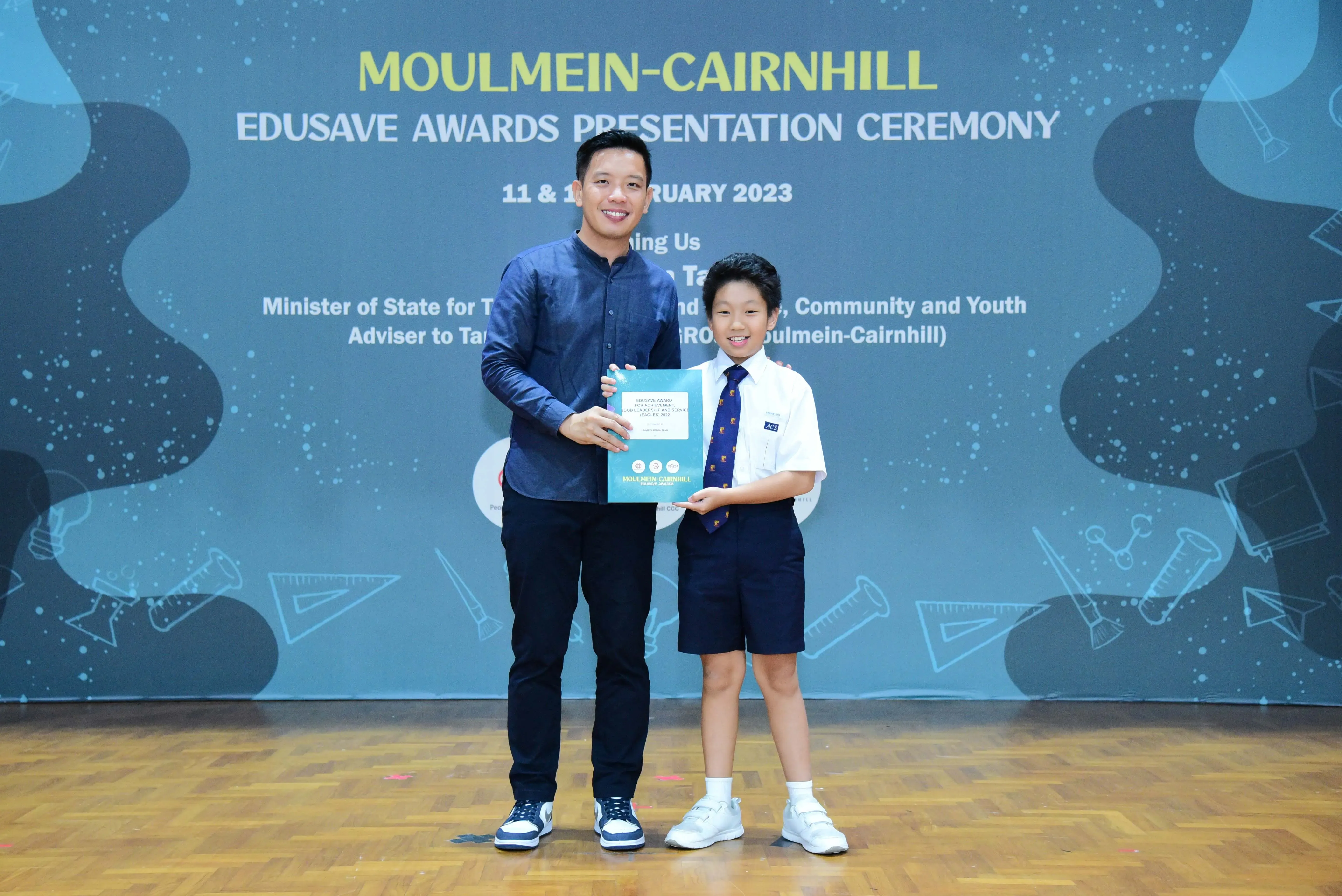 An adult and a young student in school uniform hold a certificate at the Moulmein-Cairnhill Edusave Awards Ceremony 2023.