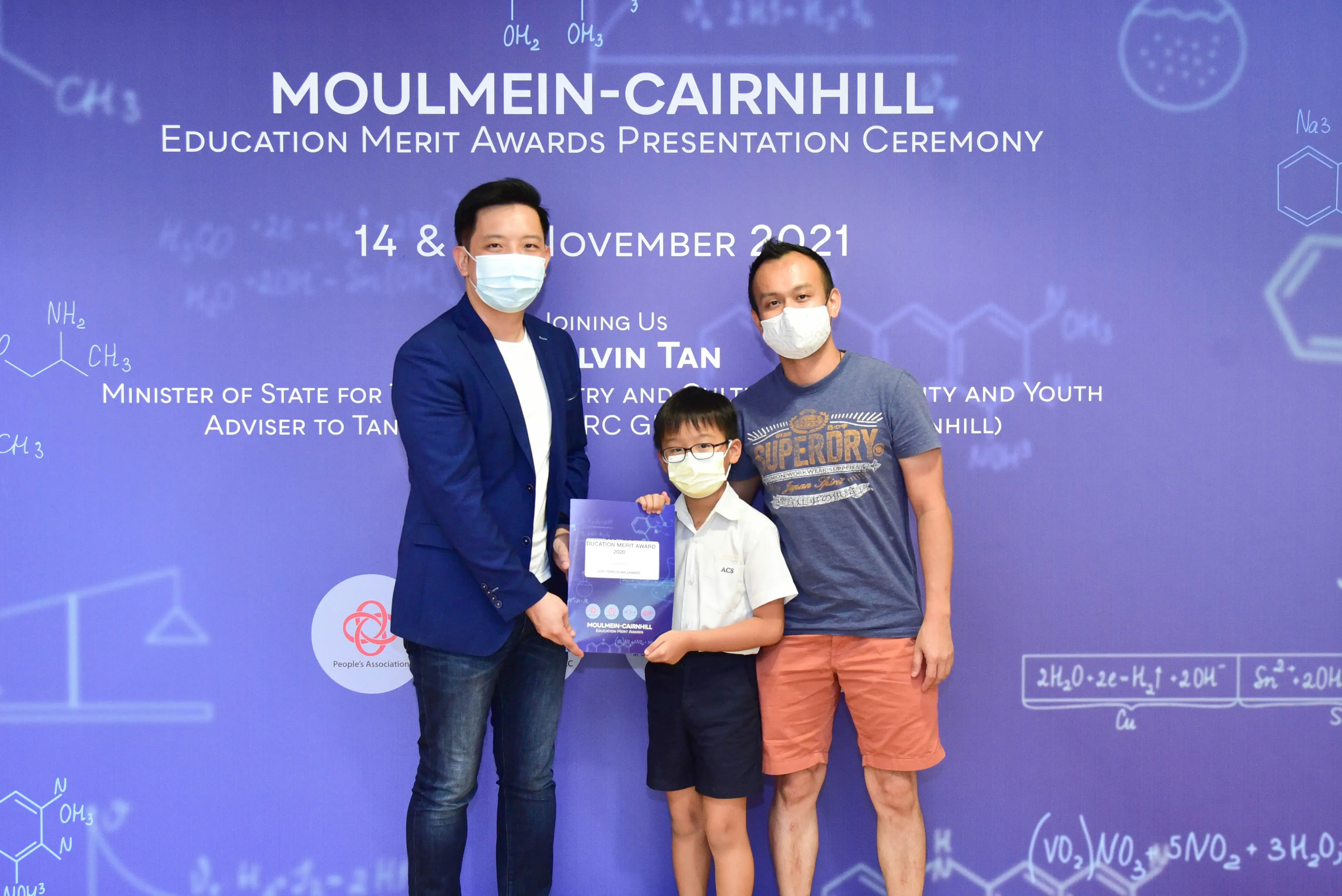 Two masked adults and a masked boy holding an award certificate at the Moulmein-Cairnhill Education Merit Awards 2021.