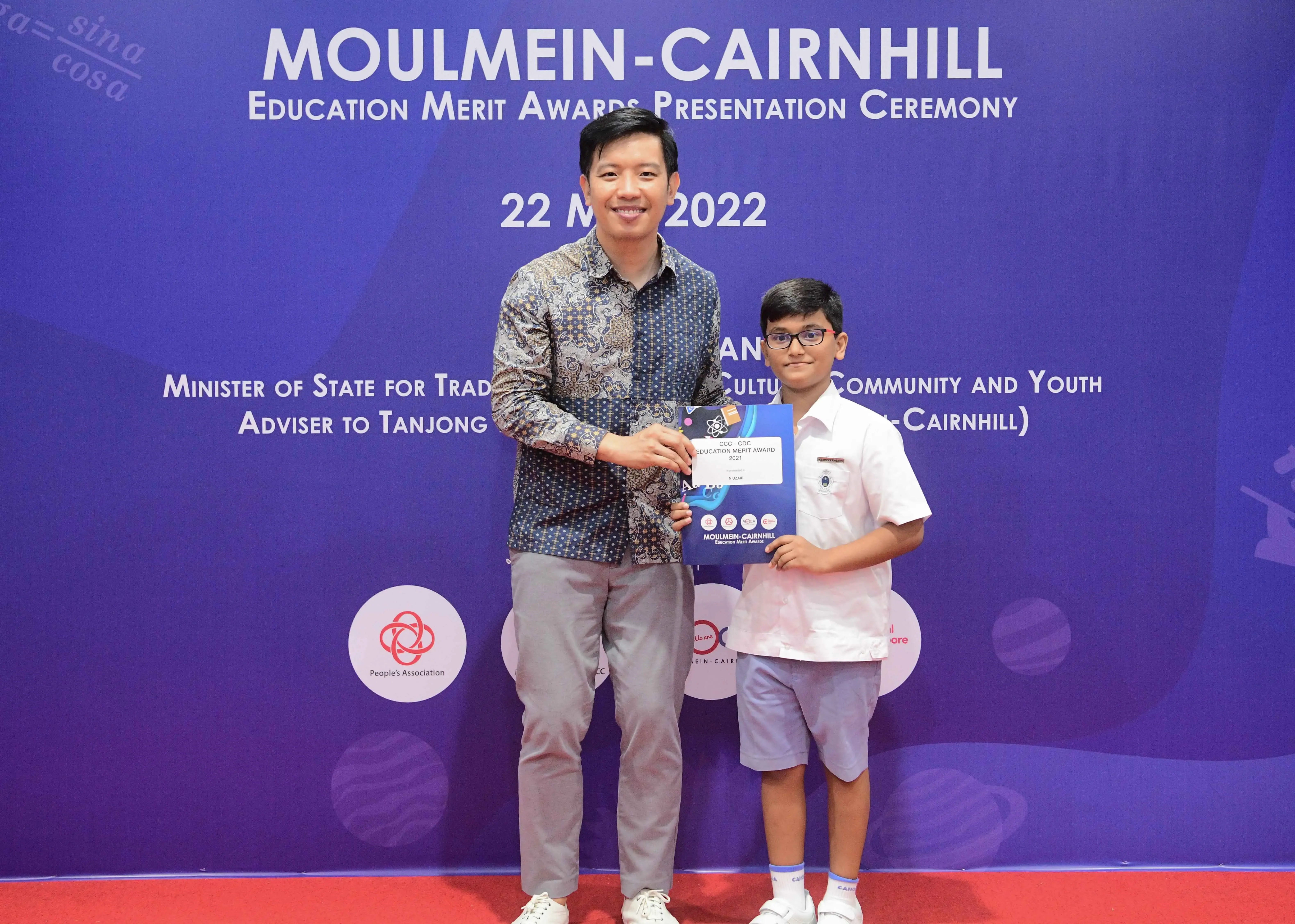 An adult man and a young boy hold a certificate at the Moulmein-Cairnhill Education Merit Awards ceremony in 2022.