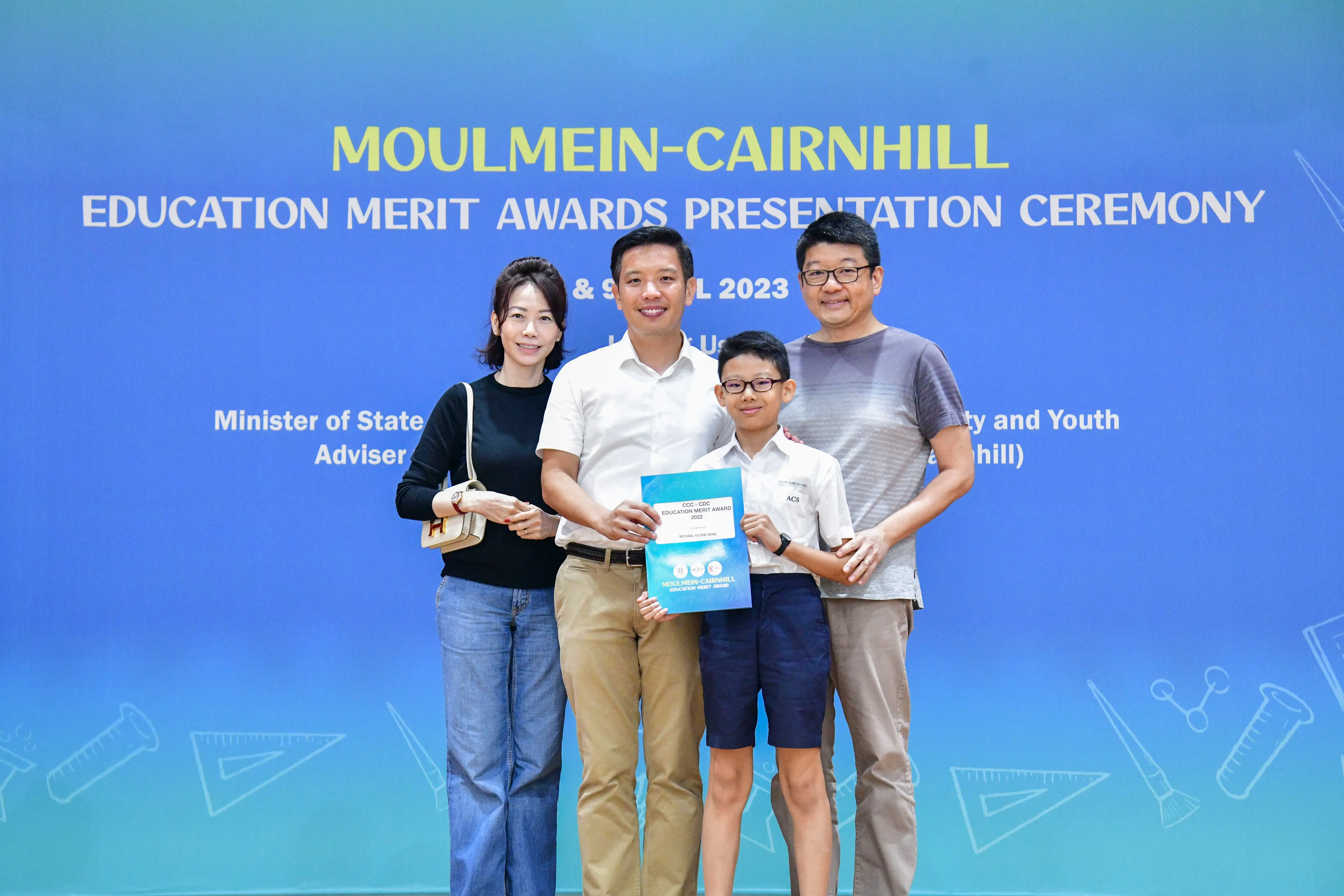 Four people pose before a blue banner for the 2023 Moulmein-Cairnhill Education Merit Awards ceremony.