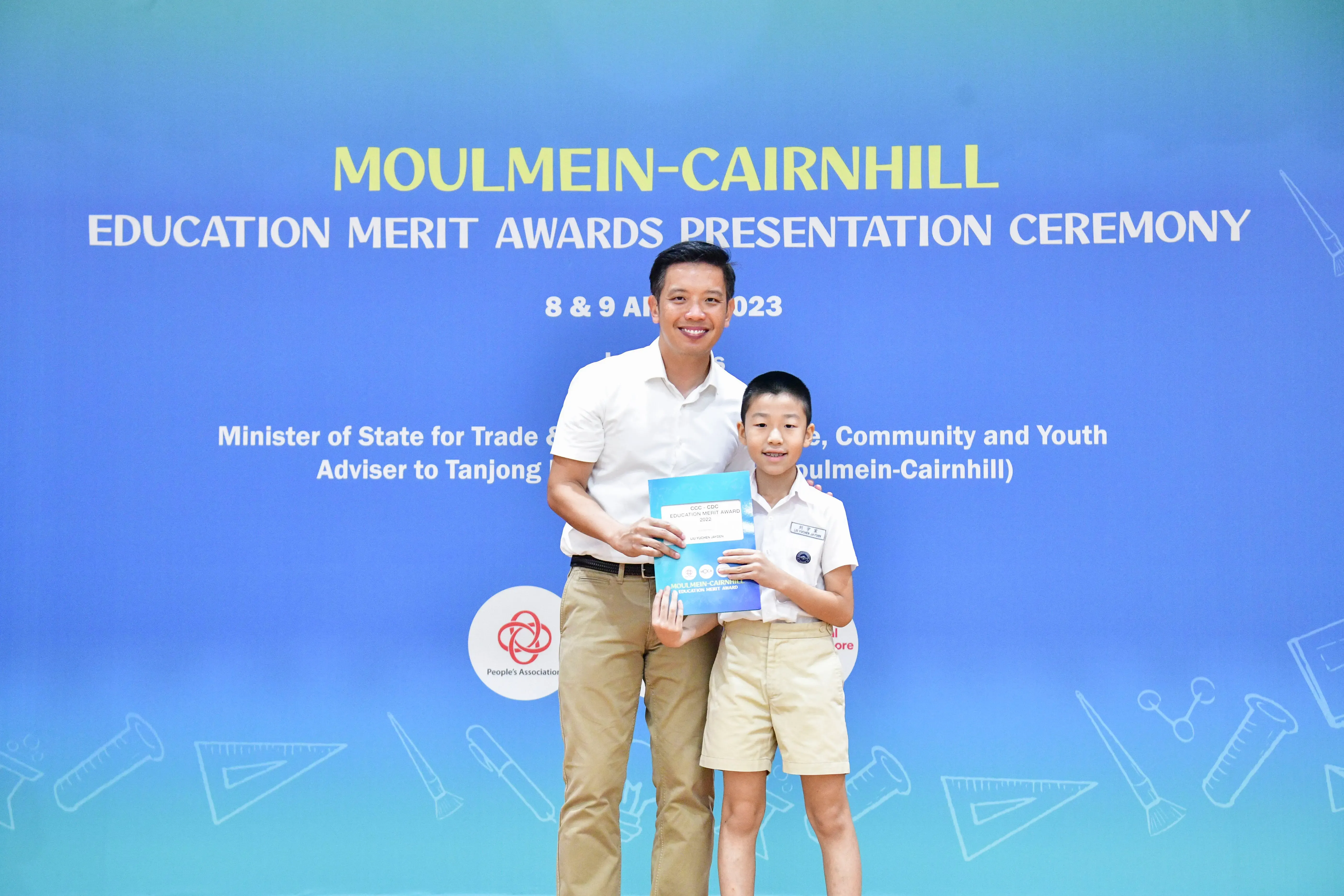 A man and a young boy hold an award certificate in front of a Moulmein-Cairnhill Education Merit Awards 2023 banner.