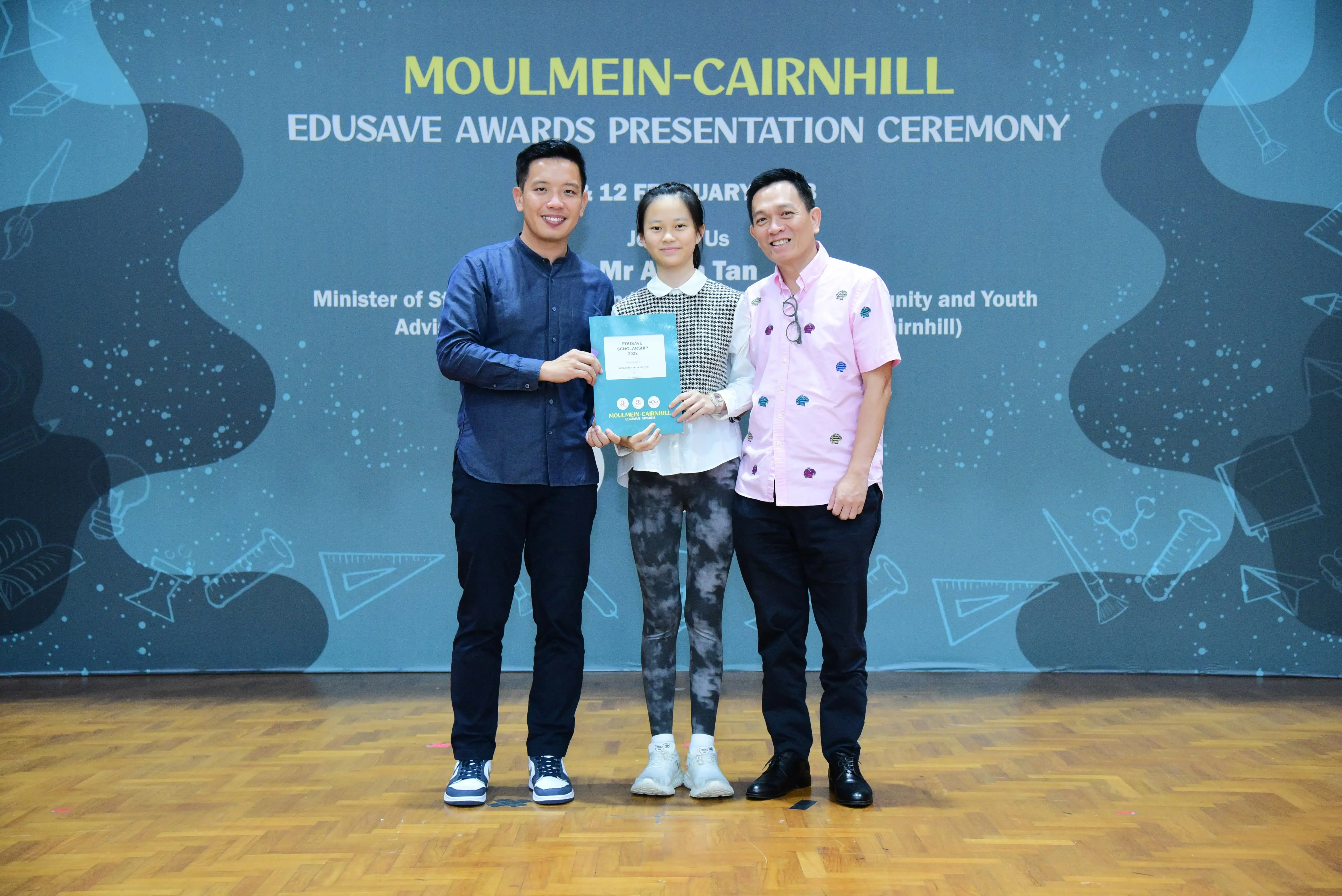Three people pose with an award folder at the Moulmein-Cairnhill Edusave Awards Presentation Ceremony backdrop.
